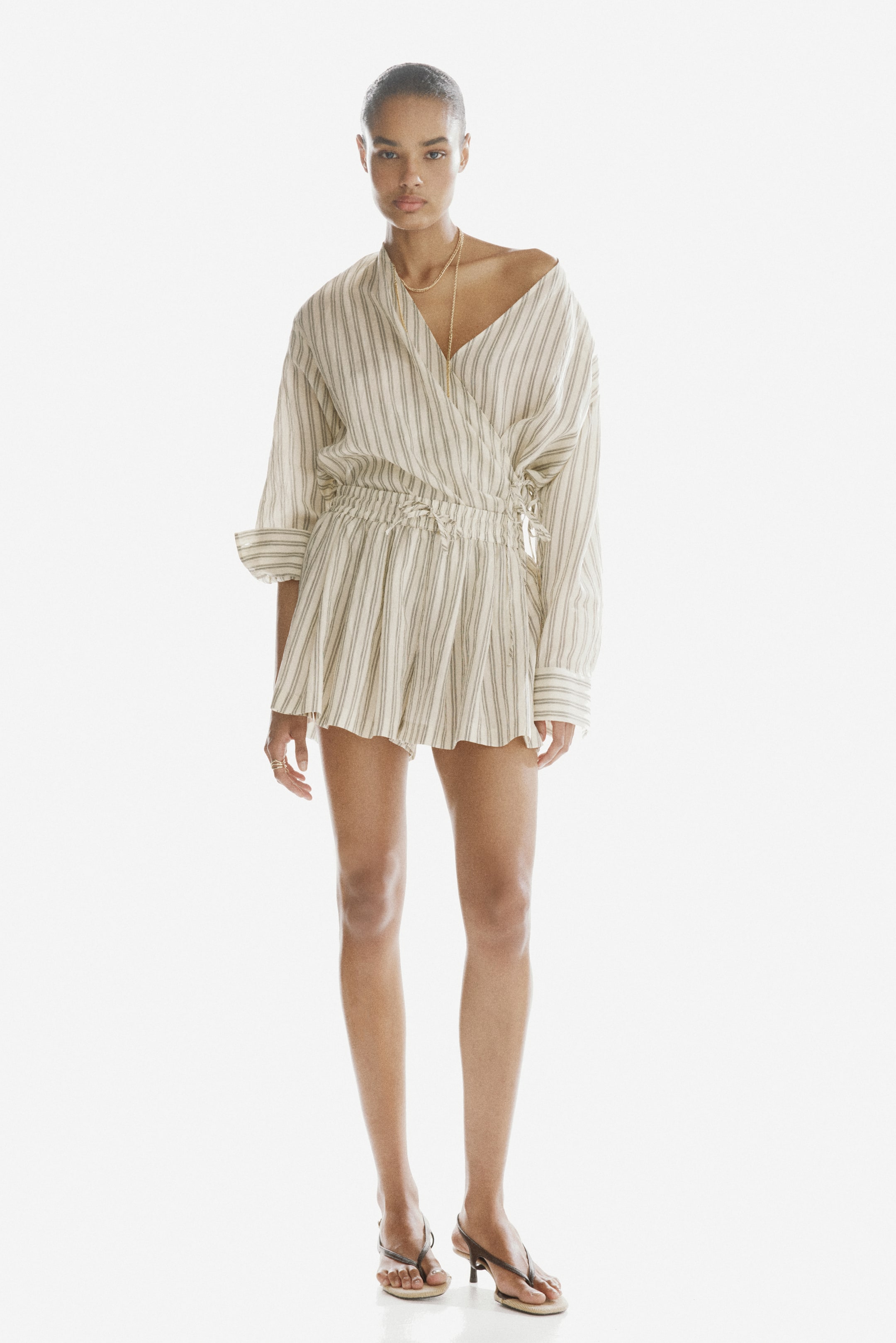 View larger image: A cream and dark striped playsuit, featuring a wrap-style V-neck top with long sleeves and cuffs, an elasticated and cinched waist with a tie detail, and loose shorts, is being worn by the model.