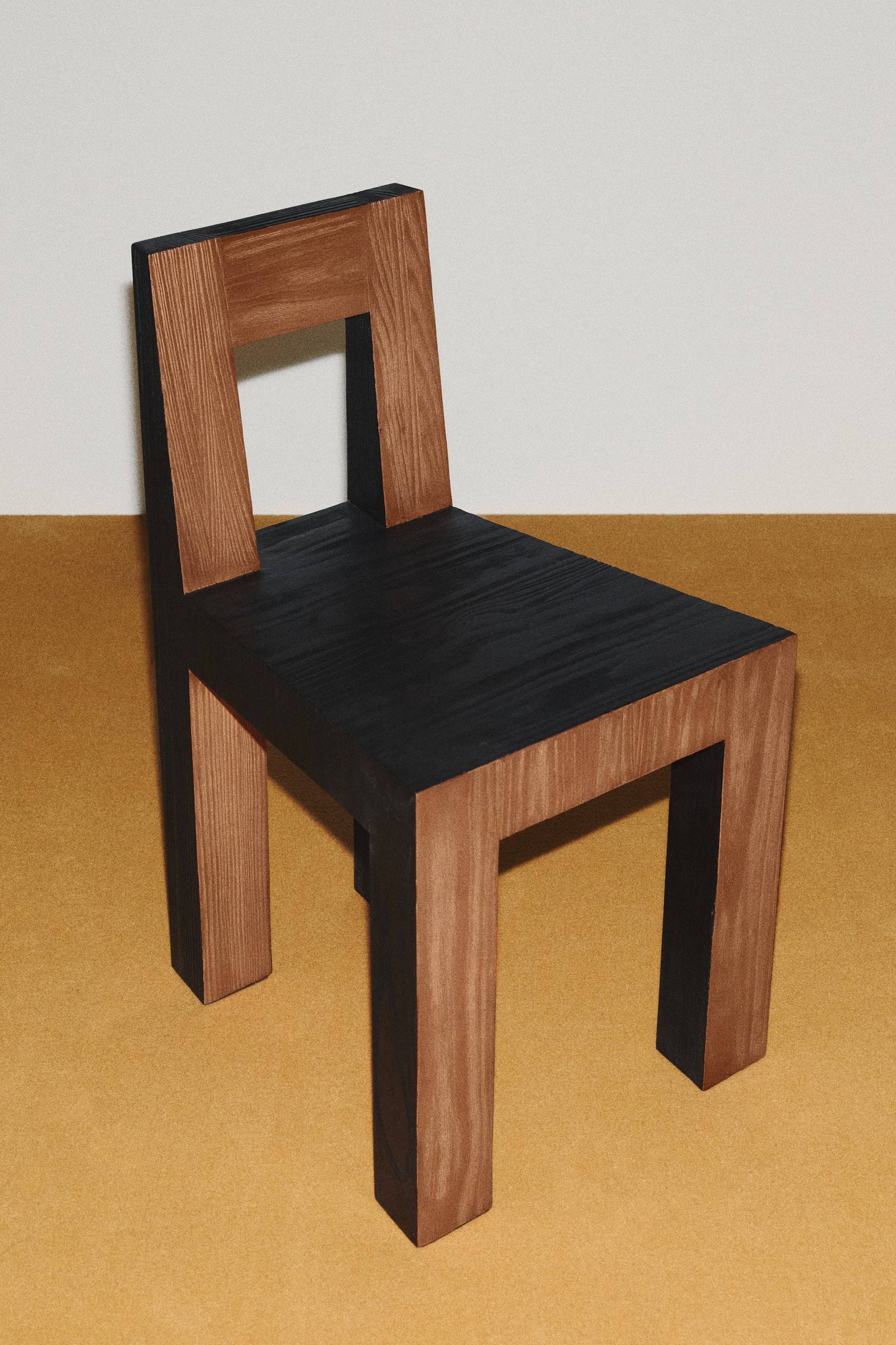 View larger image: A pinewood chair with an angular, architectural design is positioned at a three-quarter angle facing front-right. The chair features a dual-tone finish, displaying natural brown wood grain on its outer surfaces and a contrasting black finish on its flat seat and inner frames.