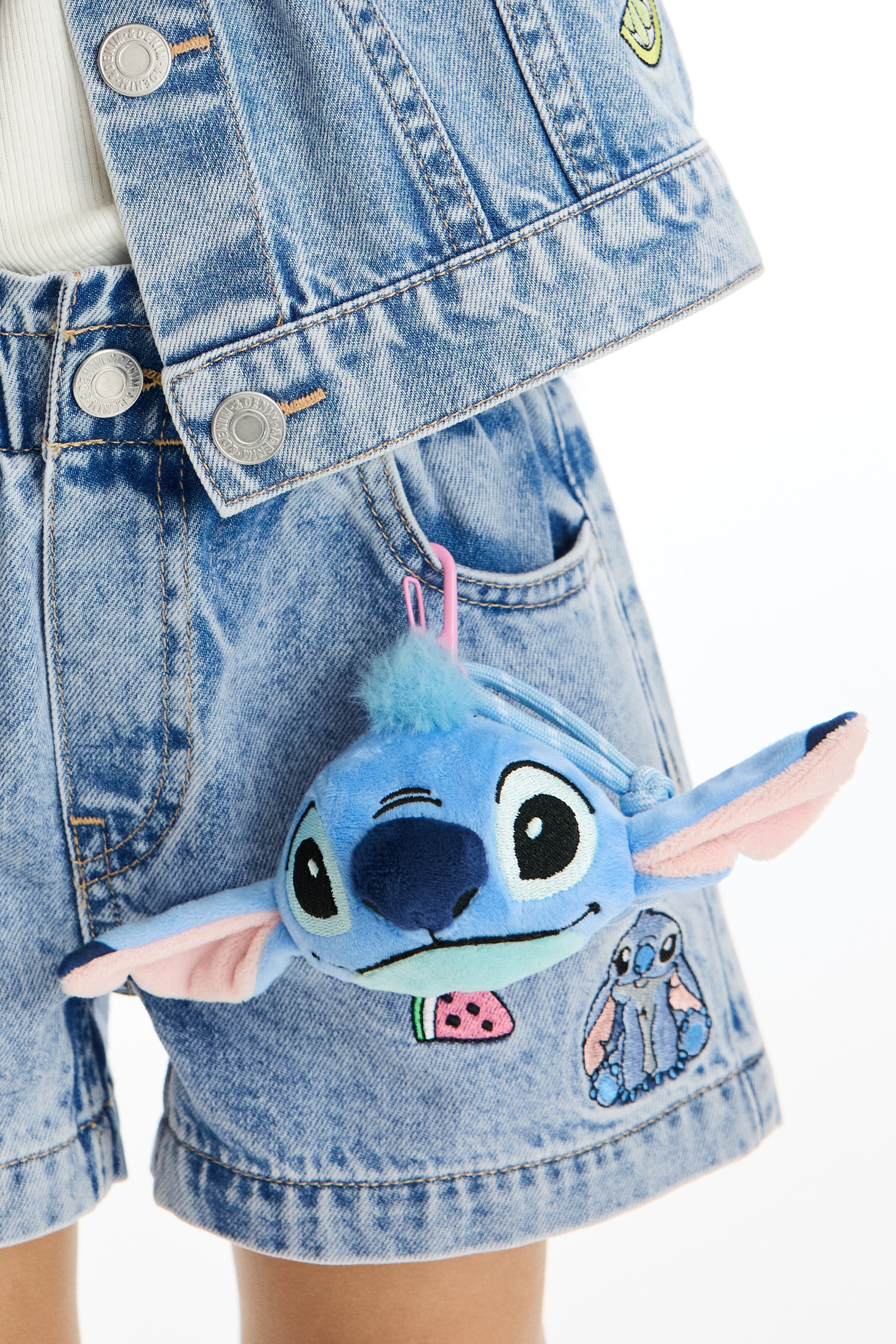 Ver imagem maior: Uma cabeça do personagem Stitch em pelúcia azul está pendurada para a frente, presa por um clipe de plástico rosa a um bolso de ganga azul claro. Apresenta grandes olhos pretos, um nariz azul escuro, orelhas internas rosa e um tufo de pelo azul claro na cabeça.