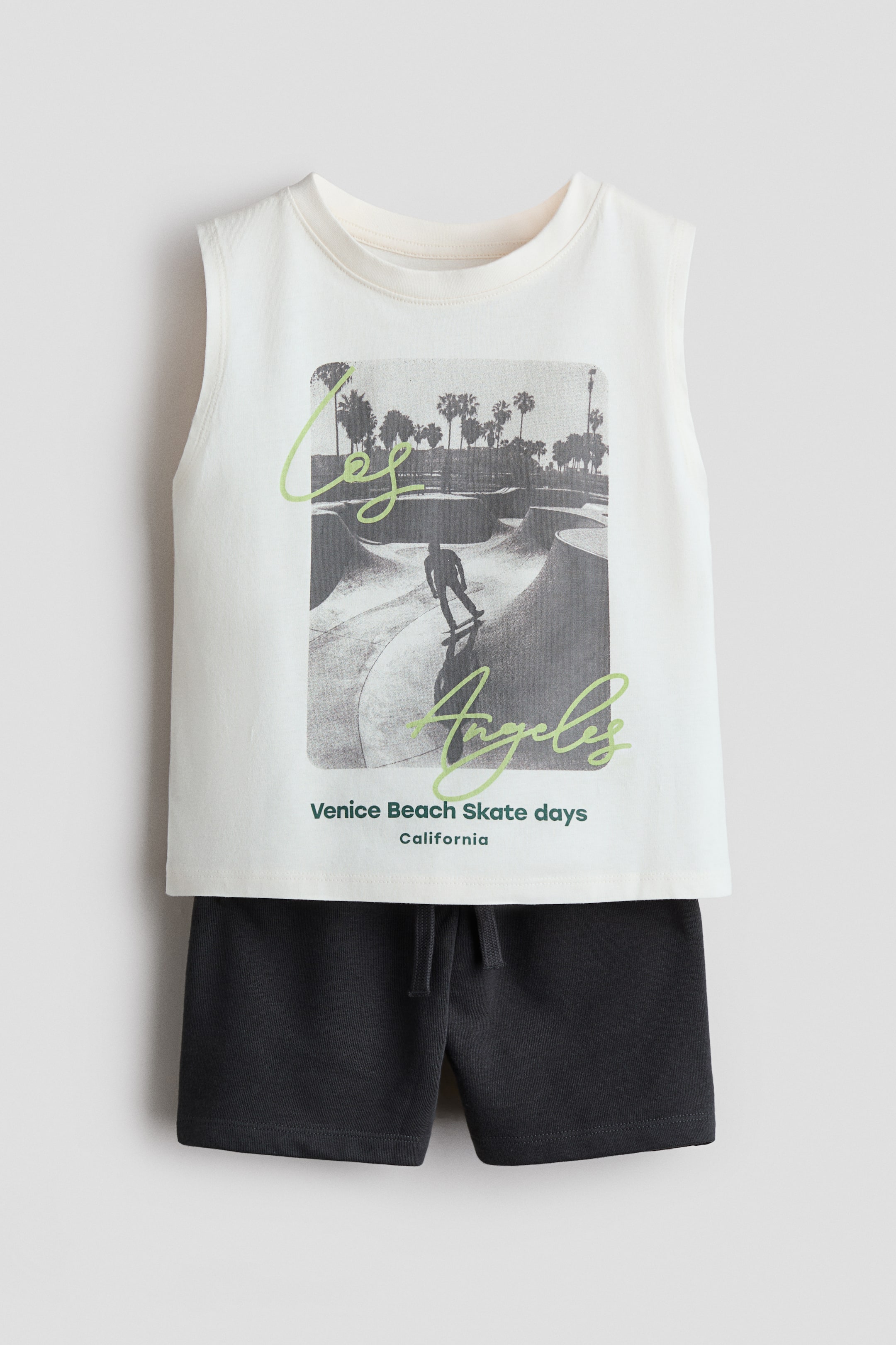 View larger image: A two-piece set, showcased from a front angle, features an off-white sleeveless top paired with dark grey shorts. The sleeveless top displays a rectangular graphic of a grayscale skate park, complete with a skateboarder and palm trees, overlaid with green script reading 'Los Angeles' and smaller text stating 'Venice Beach Skate days California.' The dark grey shorts boast an elasticated waistband with a visible drawstring tie.