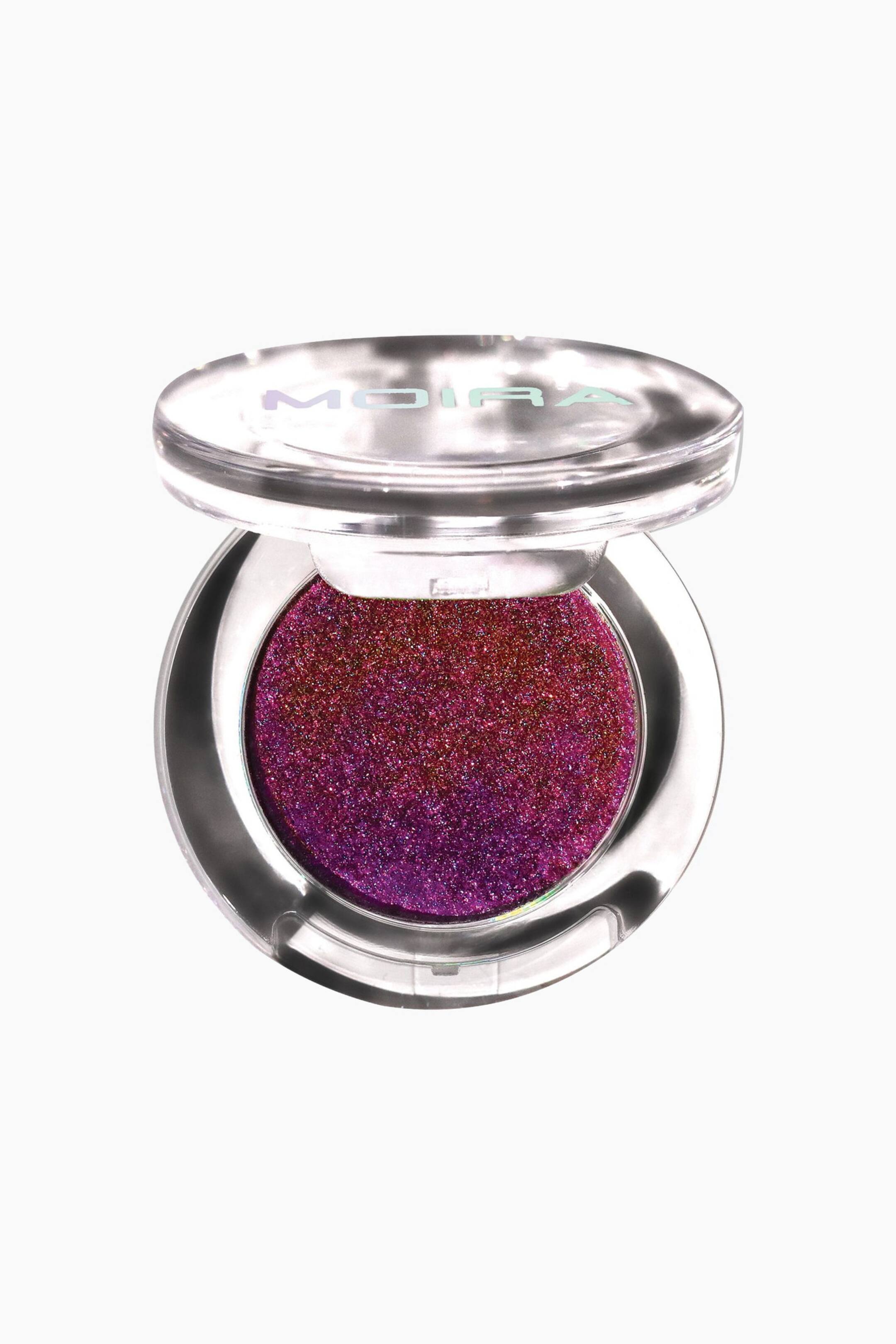 Vis større bilde: Supernova Glitz Shadow presenteres i en åpen, sirkulær, transparent akrylkompakt vendt forover. Kompakten viser en høyt teksturert, flerfarget glitterøyenskygge som går fra dyp lilla til en rødlig bronsetone, mens det baktiltede, klare lokket viser det holografiske merkenavnet «MOIRA».