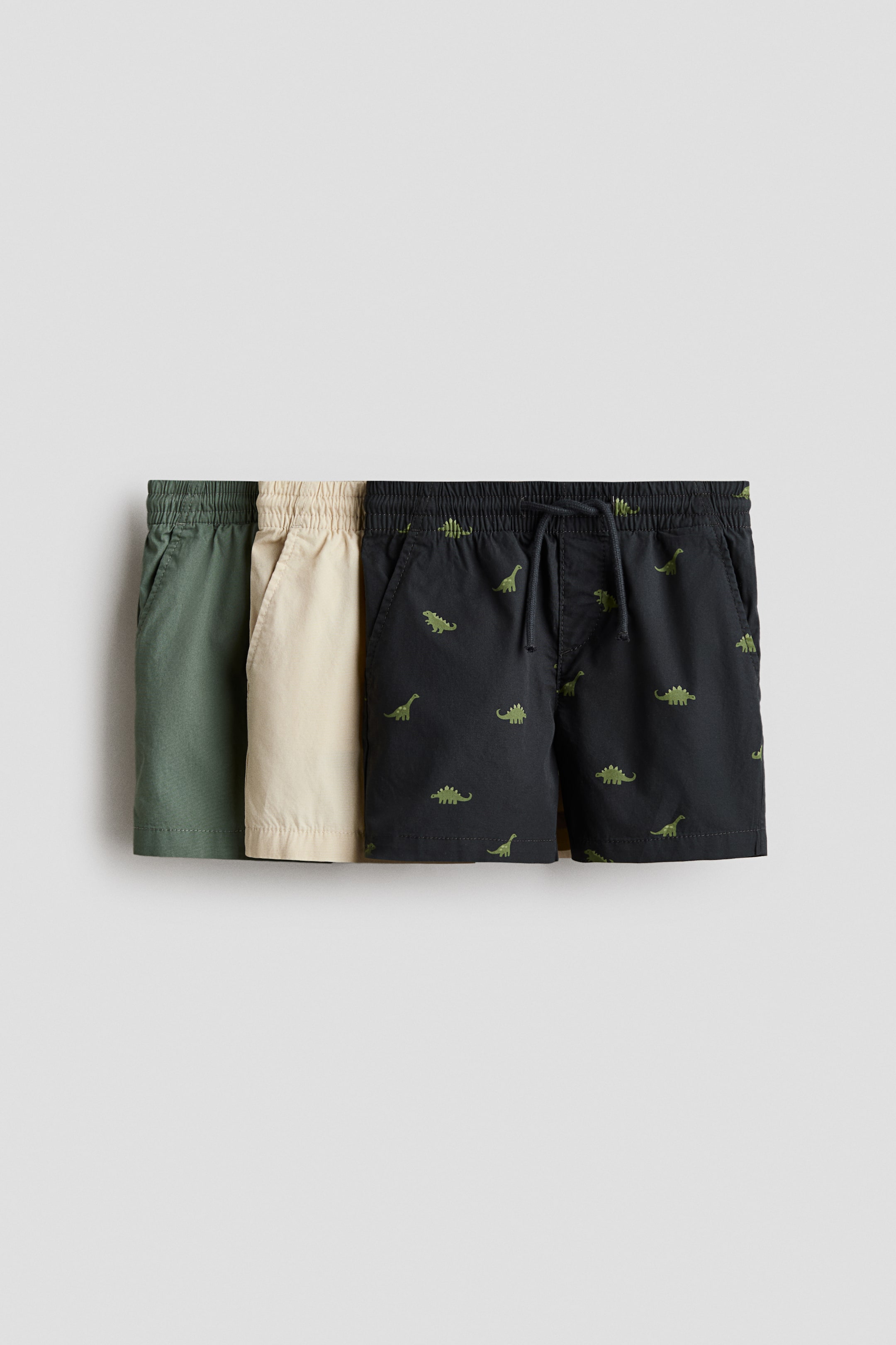 View larger image: Three pairs of shorts are laid flat, showcasing their front. They include an olive green pair, a beige pair, and a black pair adorned with a green dinosaur print. Each pair is equipped with an elasticated waistband and side pockets; the black pair also features a visible drawstring.
