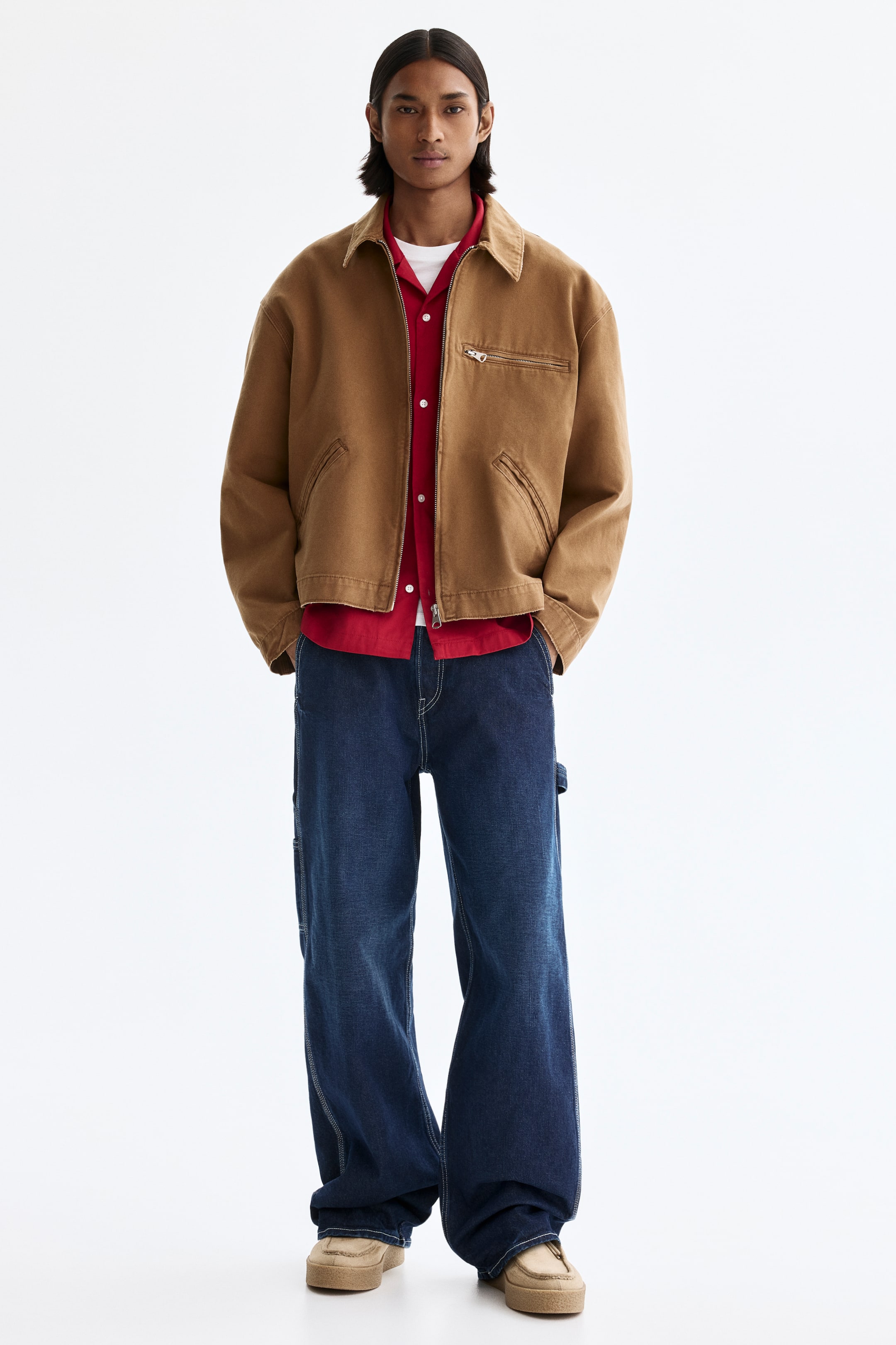 View larger image: A brown workwear-style jacket with a pointed collar, a zippered chest pocket, and two lower welt pockets is worn open to reveal a red collared shirt layered over a white crew-neck T-shirt. Dark blue wide-leg carpenter pants with contrasting stitching and a side tool loop complete the outfit, paired with tan moccasin-style shoes.
