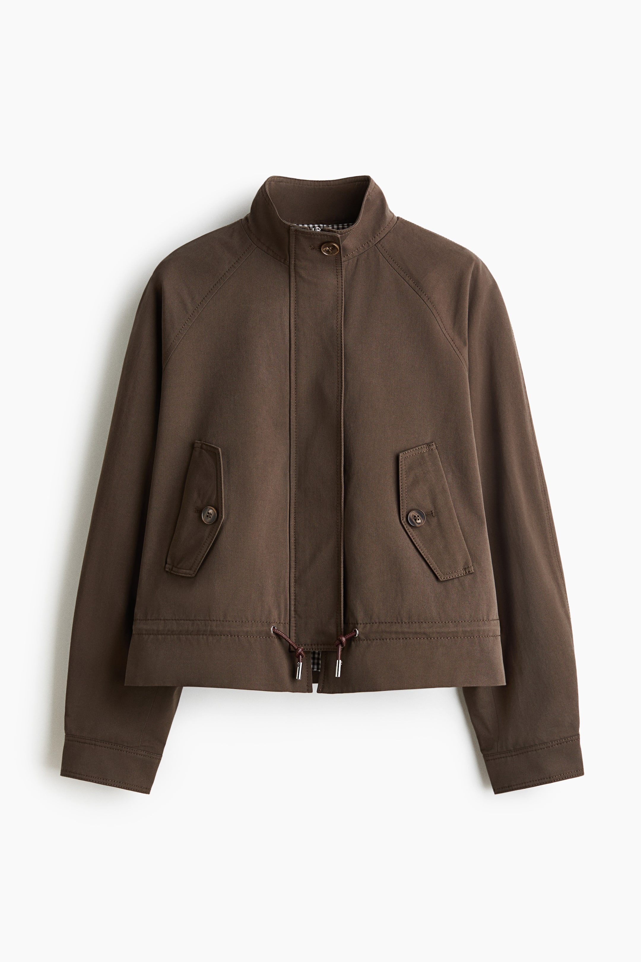 View larger image: A brown oversized cotton twill jacket is showcased from the FRONT, featuring a stand collar and a front wind flap, secured by a button at the top. It highlights raglan sleeves with buttoned cuffs, handwarmer pockets with flaps and buttons, and a drawstring above the hem, with a subtle checkered lining visible inside the collar.