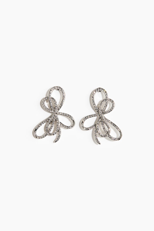 A pair of silver-toned earrings is showcasing a FRONT view, each forming an intricate, looped bow-like shape that is entirely encrusted with sparkling clear rhinestones. - 1