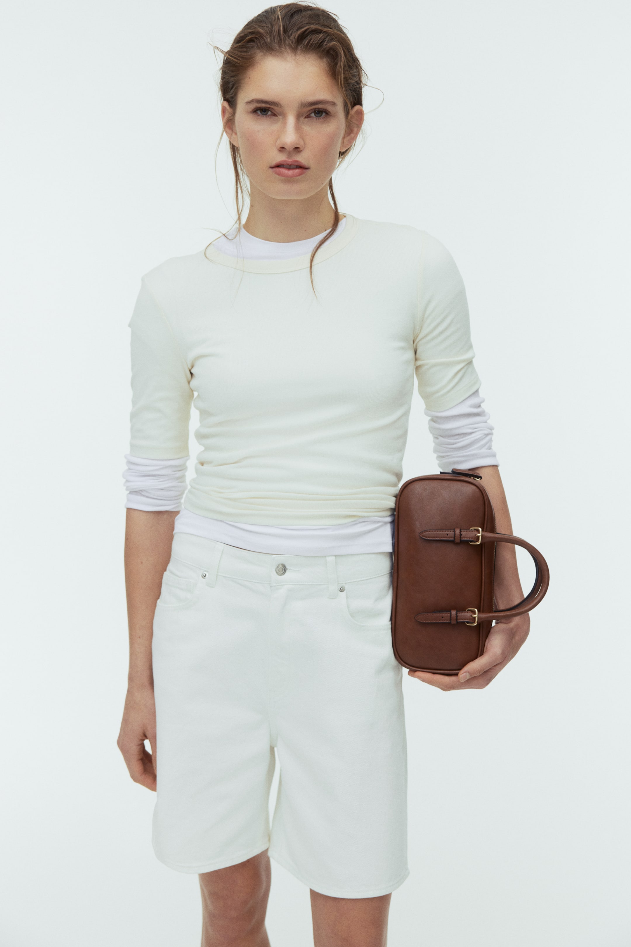 View larger image: A brown elongated bag is held upright in a hand, its front facing the camera. It features a single curved handle and two vertical decorative straps with gold-toned buckles.