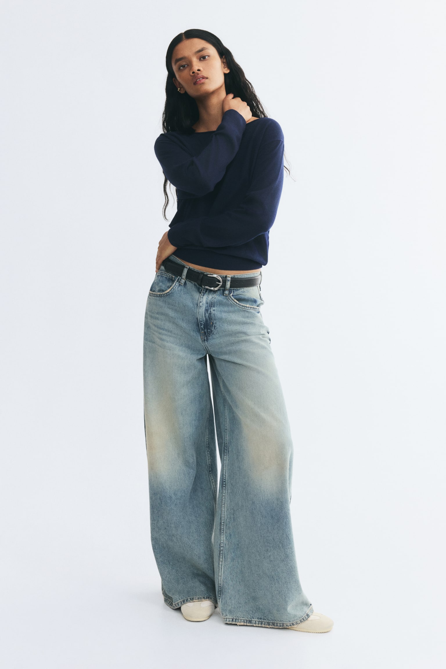 A dark blue knit sweater featuring a wide off-the-shoulder neckline, dropped shoulders, and ribbed cuffs is being worn by the model. The sweater is styled with wide-leg, light-wash denim jeans and a black belt, with the model resting one hand against their neck. - 1
