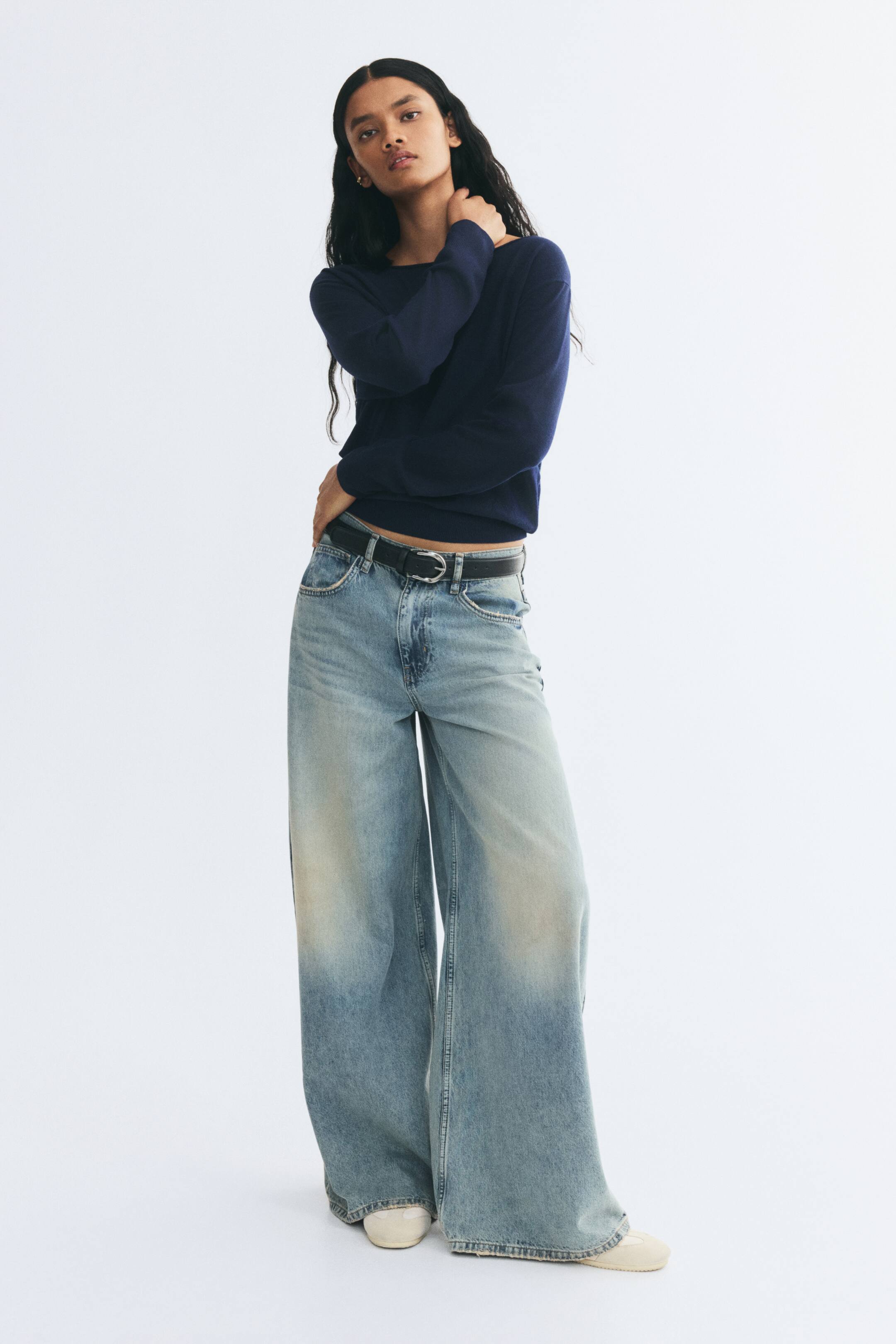 View larger image: A dark blue knit sweater featuring a wide off-the-shoulder neckline, dropped shoulders, and ribbed cuffs is being worn by the model. The sweater is styled with wide-leg, light-wash denim jeans and a black belt, with the model resting one hand against their neck.
