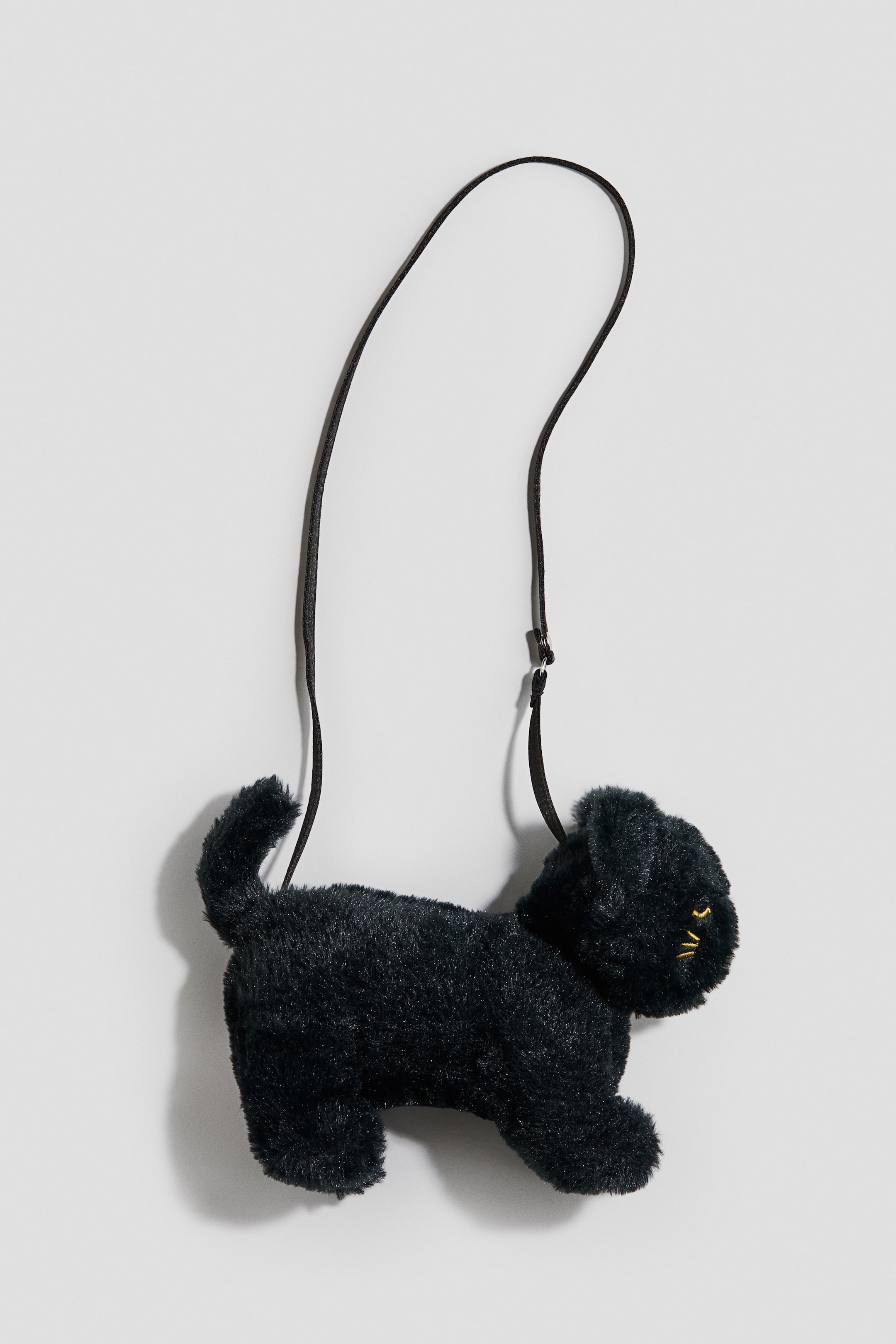 View larger image: A black plush shoulder bag shaped like a cat is hanging in profile facing right, showcasing a fuzzy texture and an upright tail. The bag is displaying yellow embroidered eyes and whiskers, with a thin black strap looping upward.