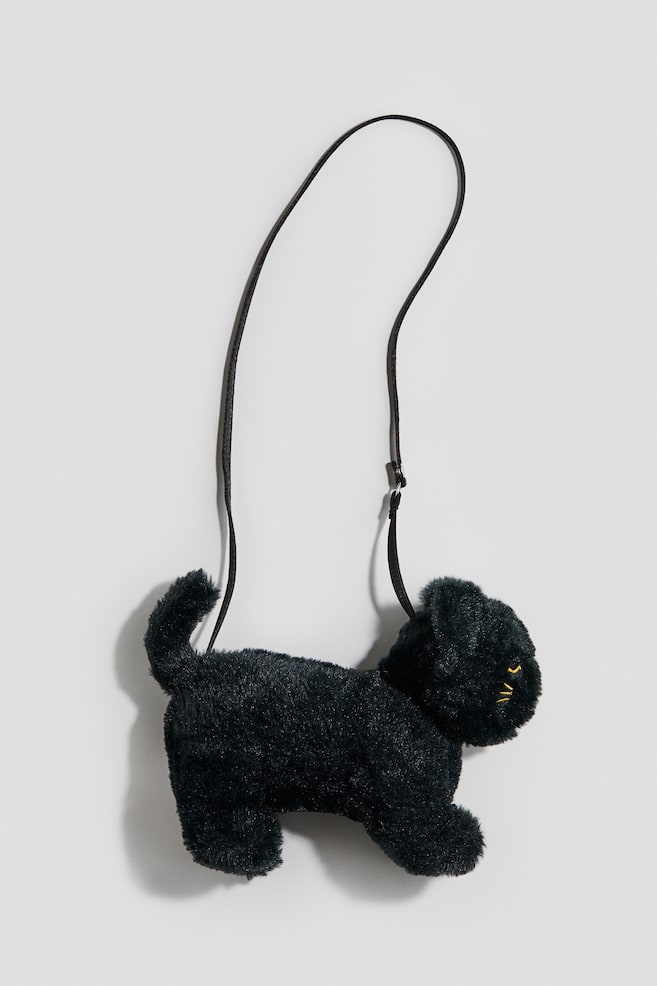Un sac bandoulière noir en peluche, en forme de chat, est suspendu de profil, face à droite, mettant en valeur une texture duveteuse et une queue dressée. Le sac présente des yeux et des moustaches brodés jaunes, avec une fine bandoulière noire qui remonte en boucle. - 1