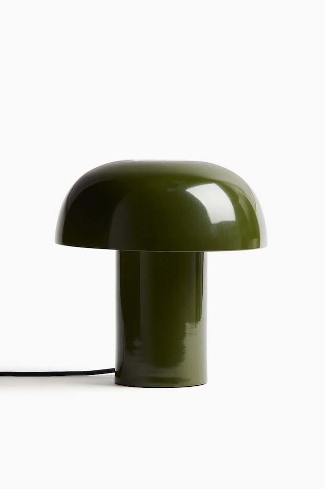 Eine moderne Tischlampe wird von vorne gezeigt, mit einer glänzenden Metallkonstruktion in dunklem Olivgrün und einer weichen, pilzförmigen Form. Ein mit Stoff überzogenes schwarzes Kabel erstreckt sich von ihrem zylindrischen Sockel. - 1