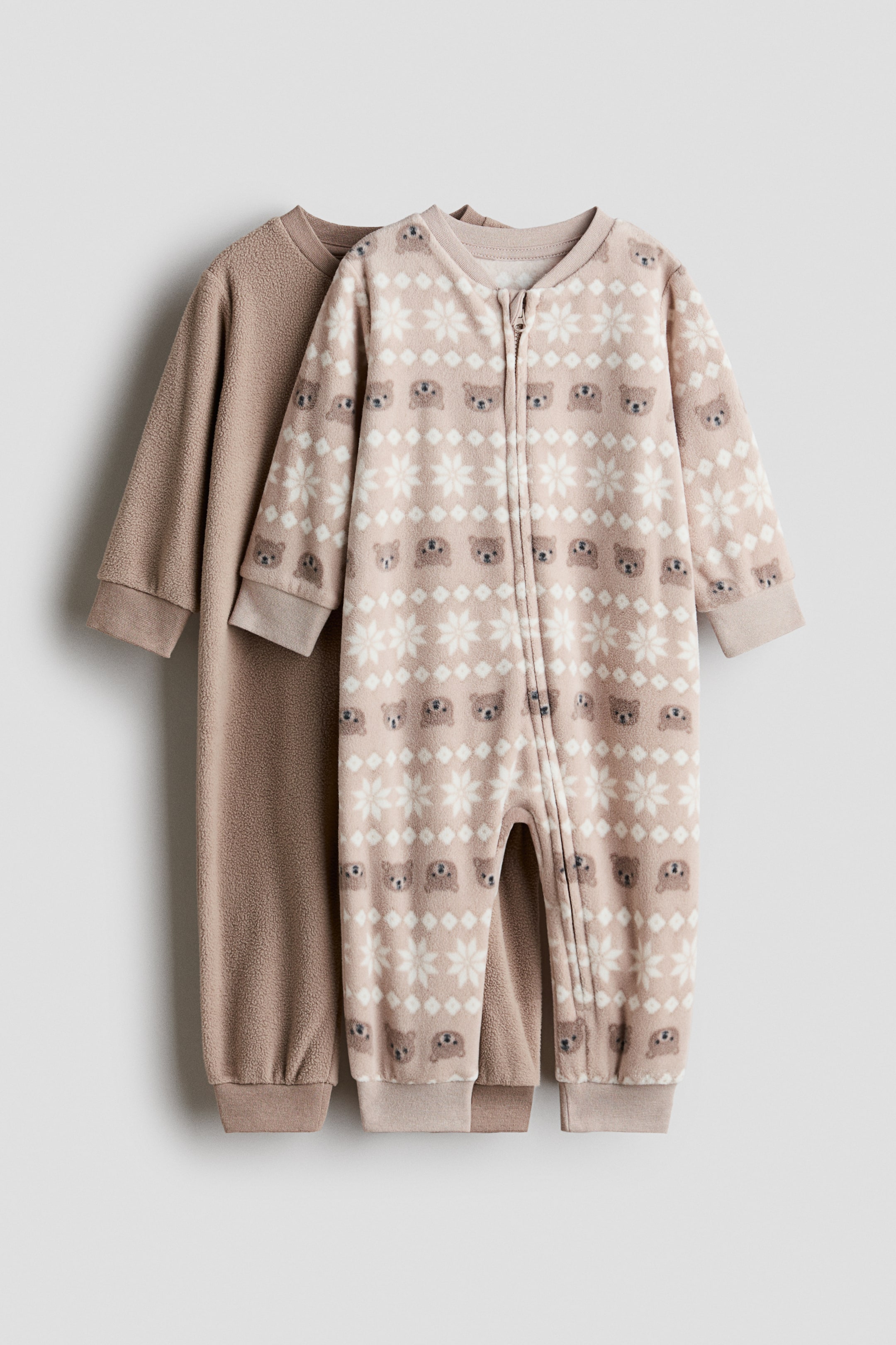 View larger image: Two long-sleeved fleece pyjama jumpsuits are hanging front-facing, with the front jumpsuit showing a pattern of white snowflakes and small brown bear faces on a light beige background. The front jumpsuit features a zip running from the round neckline down one leg, while the second jumpsuit at the back is a solid light brown colour, both designed with jersey trim at the cuffs and hems.