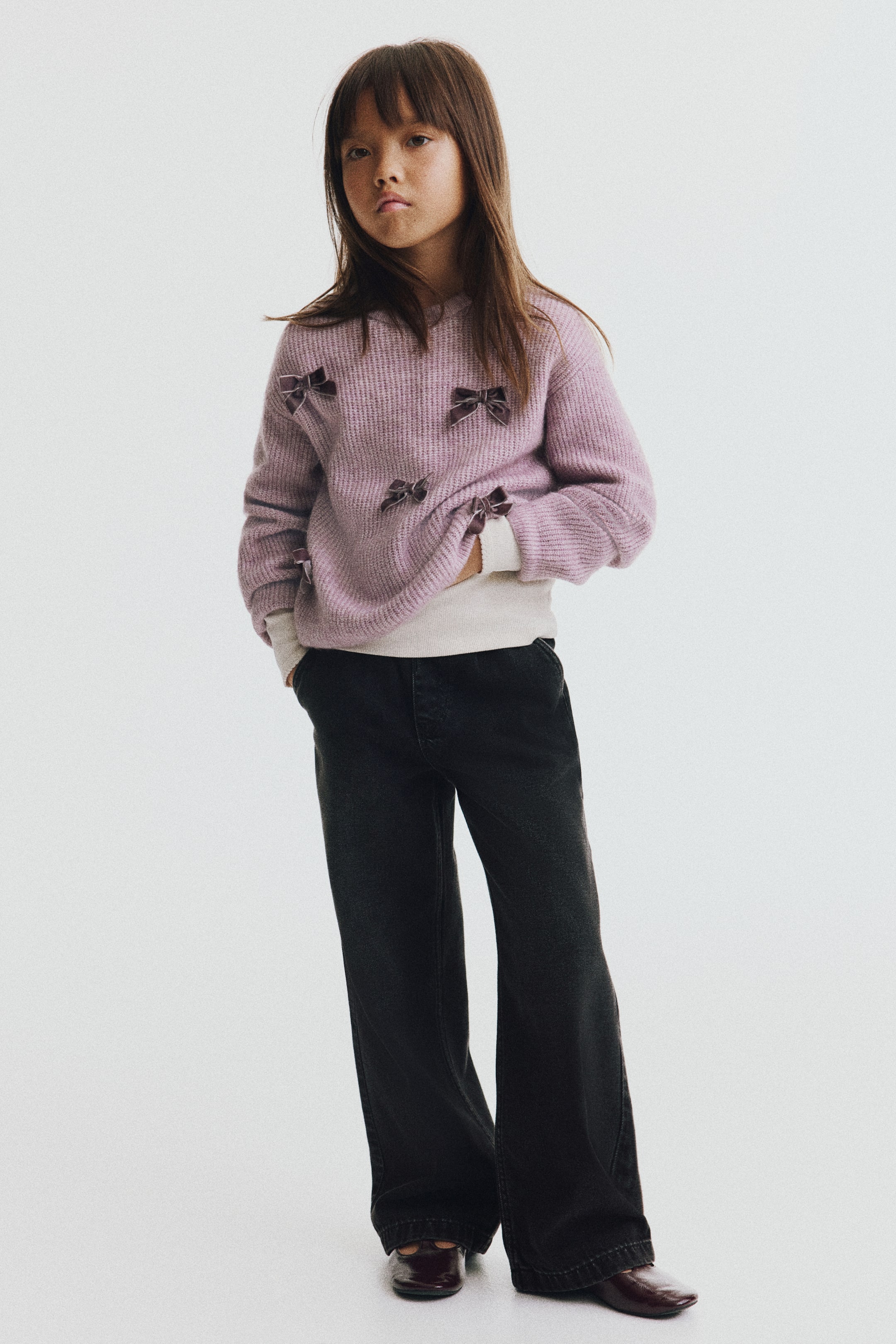 View larger image: Black cotton denim jeans, shown from the front, feature a wide-leg silhouette and diagonal side pockets. The relaxed-fit jeans are styled with a cream top and a lilac knit sweater to highlight their casual drape.