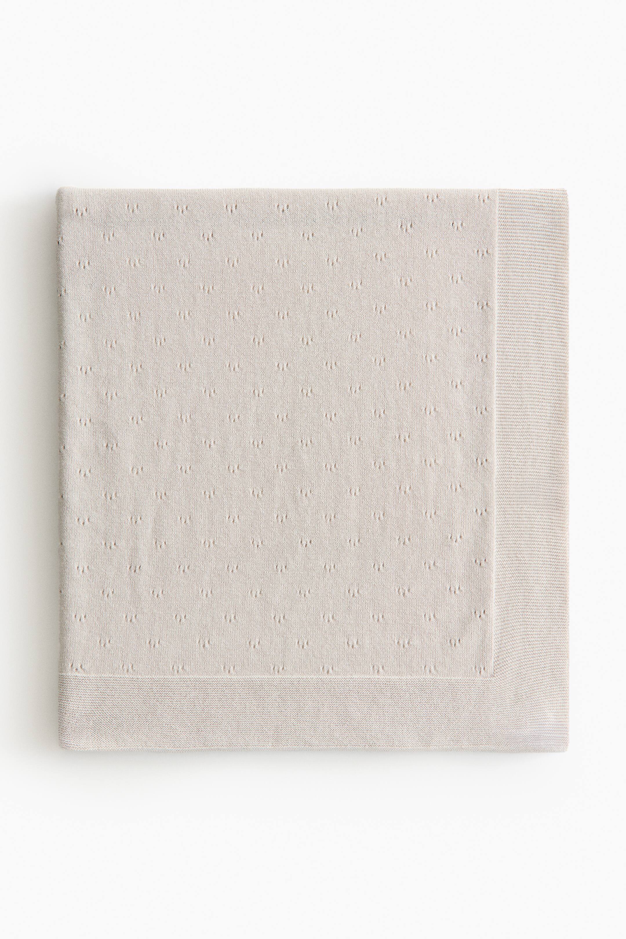 拡大画像を表示: ライトなニュートラルカラーのベビーブランケットは、ソフトなコットンのポワンテールニットを特徴とし、メインボディ全体に小さな浮き上がったドットのテクスチャパターンを見せています。すべての見える縁には控えめなプレーンニットのボーダーが施され、長方形の形にきれいに折りたたまれ、正面から平らに置かれています。