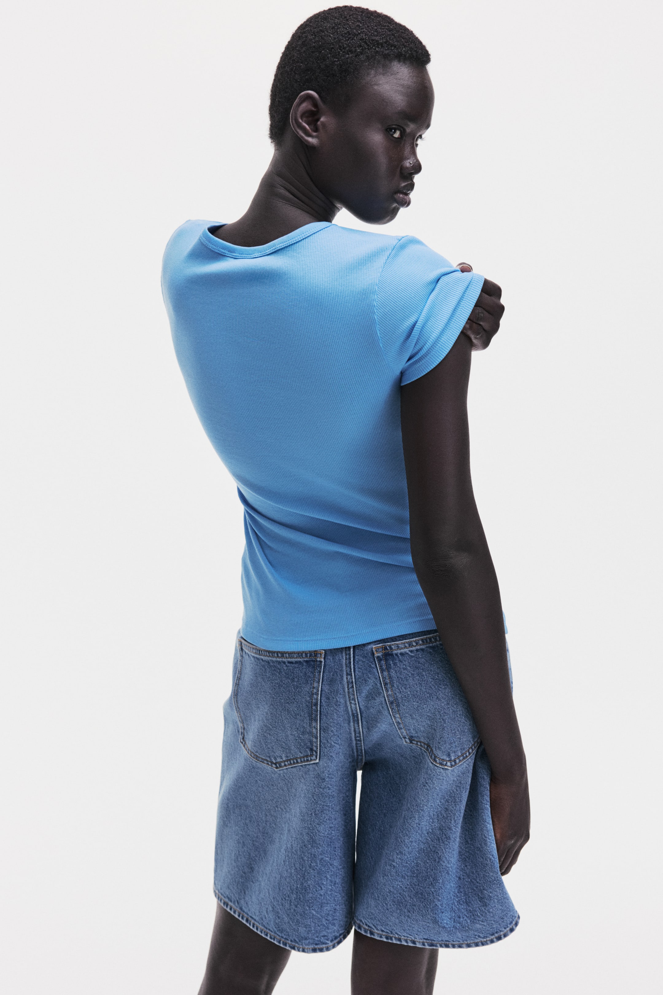 View larger image: A blue ribbed short-sleeved T-shirt is shown from the back, angled slightly to the right. It features a ribbed crew neck and a fitted silhouette that ends at the upper hips.
