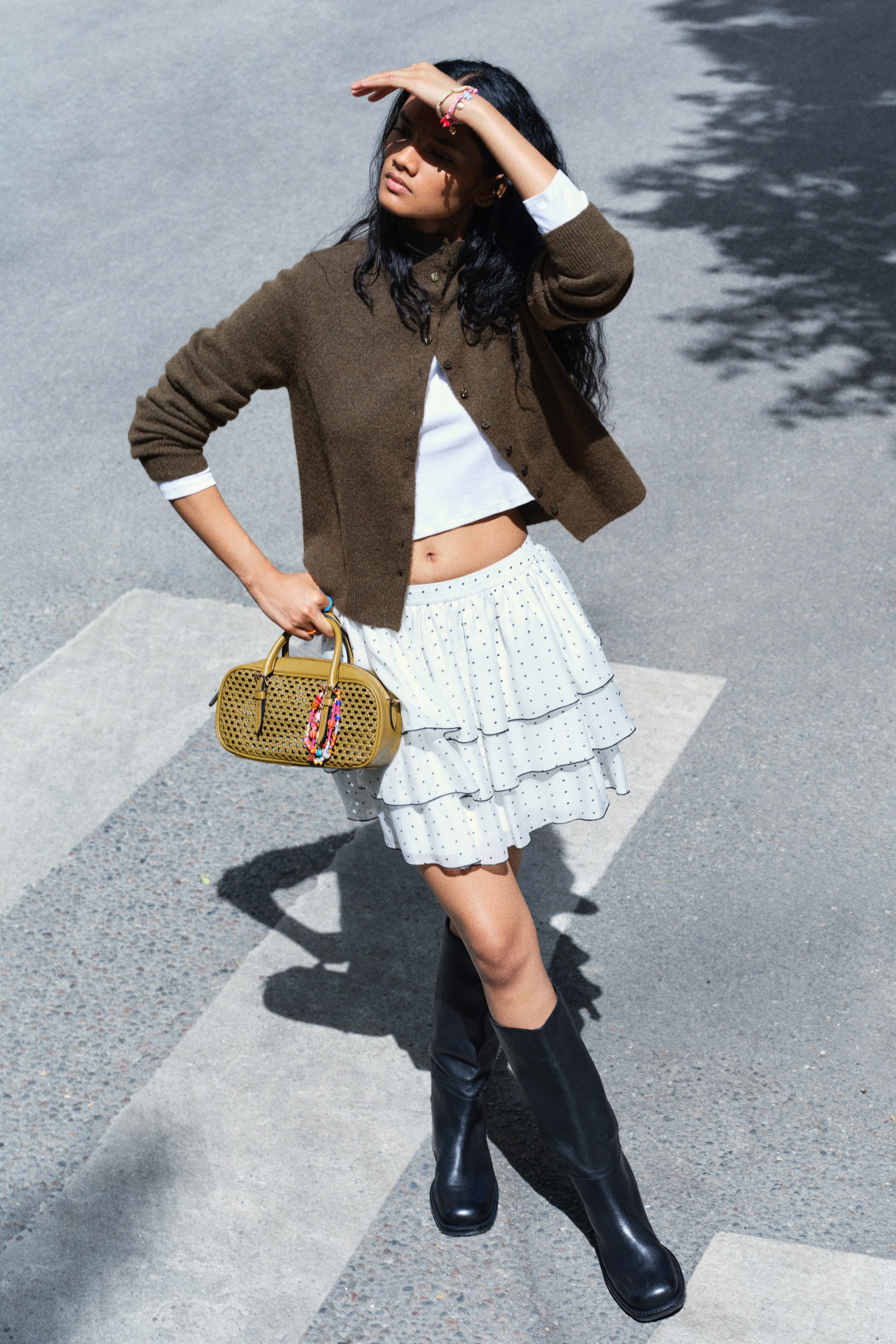 View larger image: The model is wearing a brown knitted cardigan and a tiered white polka-dot mini skirt, and is carrying a small olive-green woven handbag. The cardigan is styled open over a white crop top and paired with black leather knee-high boots.