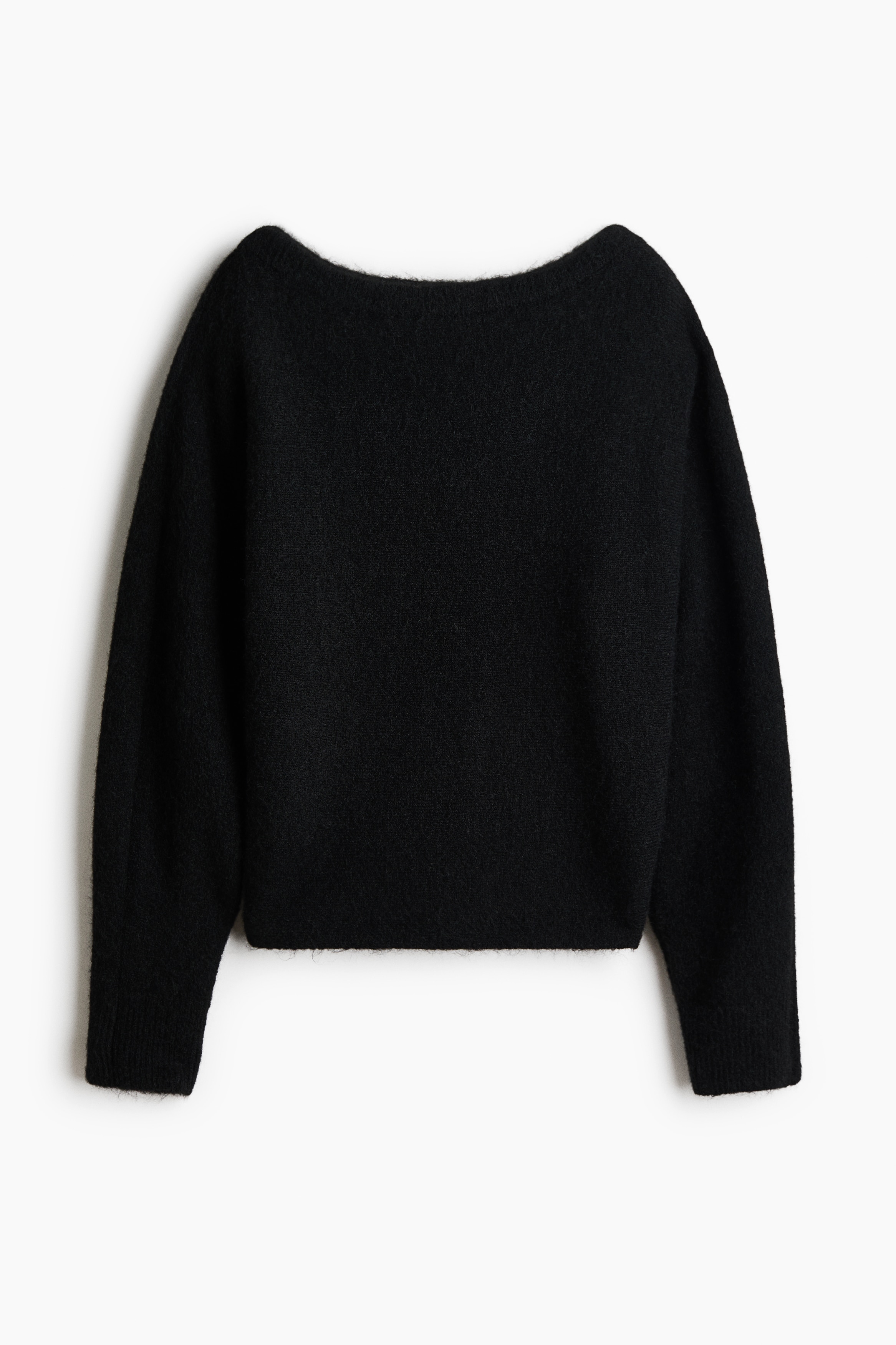 A black jumper in a soft brushed knit is being worn by the model over a layered white top and black trousers. The jumper features a wide boat neck and long sleeves, with the model resting one hand near their hip.
