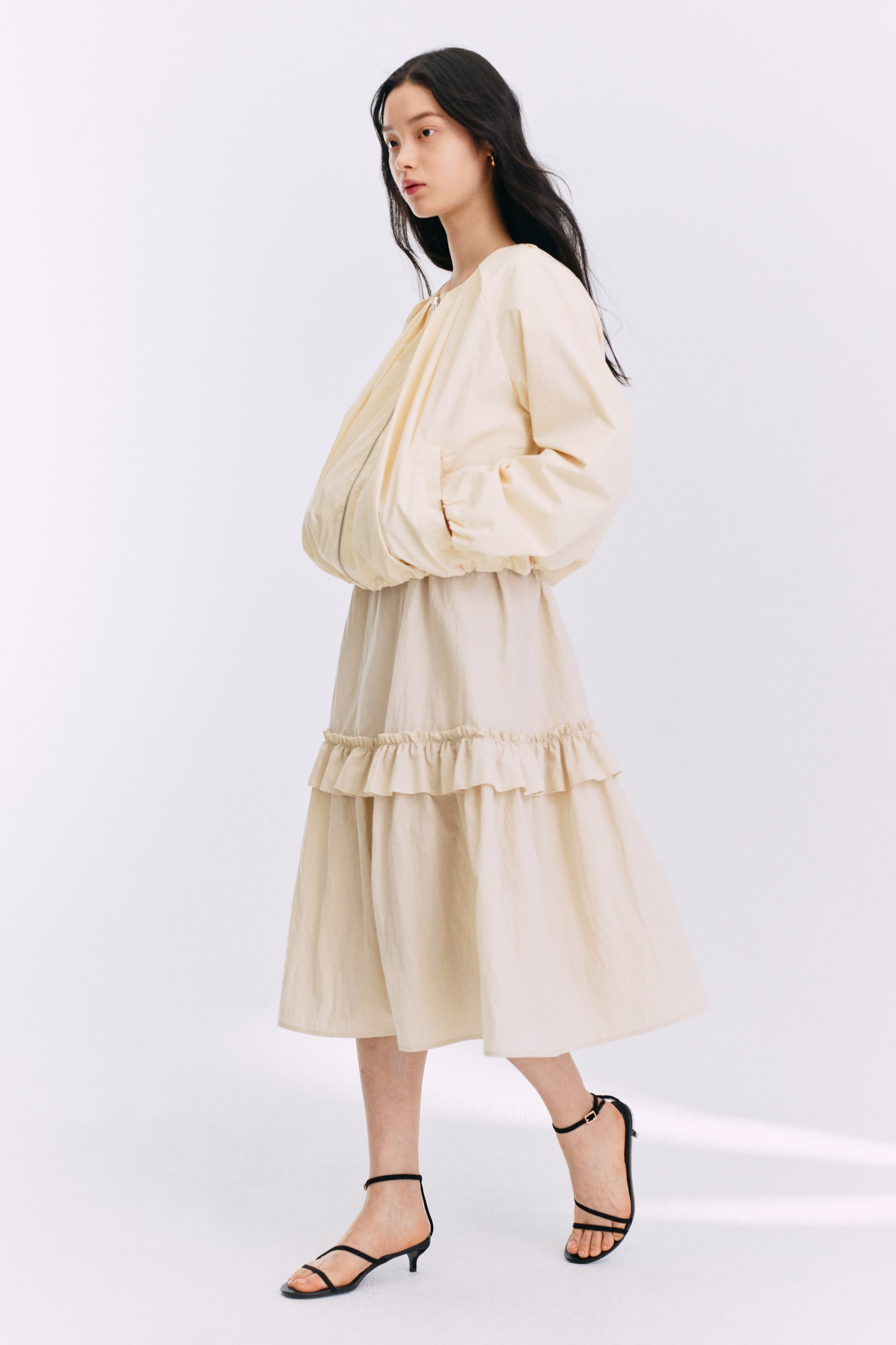 拡大画像を表示: クリーム色のドレスは、前面にジッパーが付いたボリュームのあるボンバースタイルのトップと、ギャザー入りのウエストが特徴で、フリルの縫い目があるティアードミディスカートに繋がっています。わずかに横からのアングルでご紹介します。