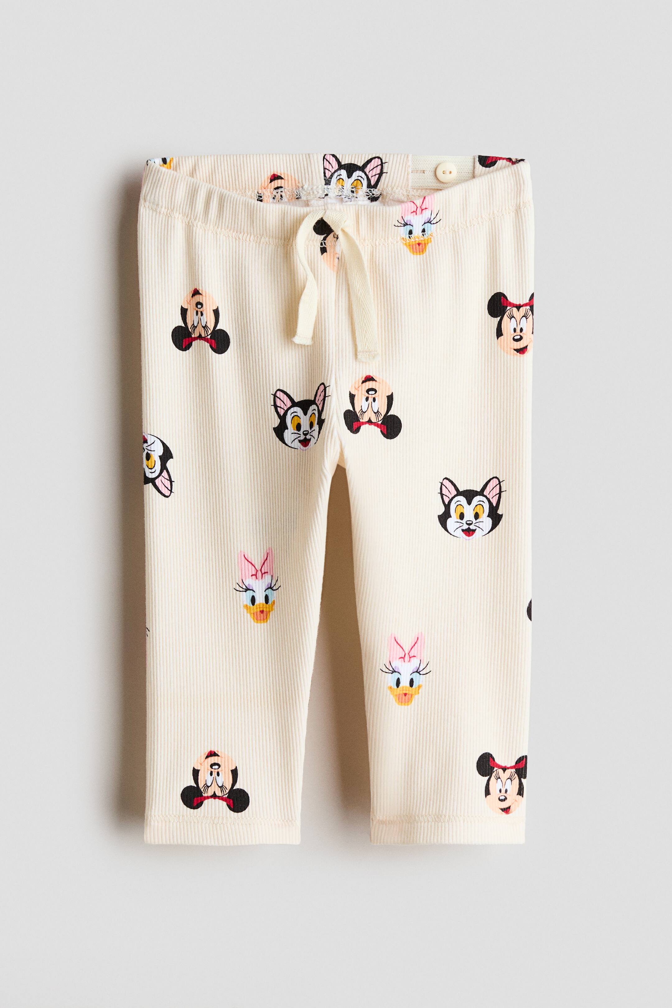 View larger image: Cream ribbed baby trousers are shown from the front, highlighting an elastic waistband with an adjustable drawstring and internal button adjusters, along with a full print of Minnie Mouse, Daisy Duck, and Figaro faces.