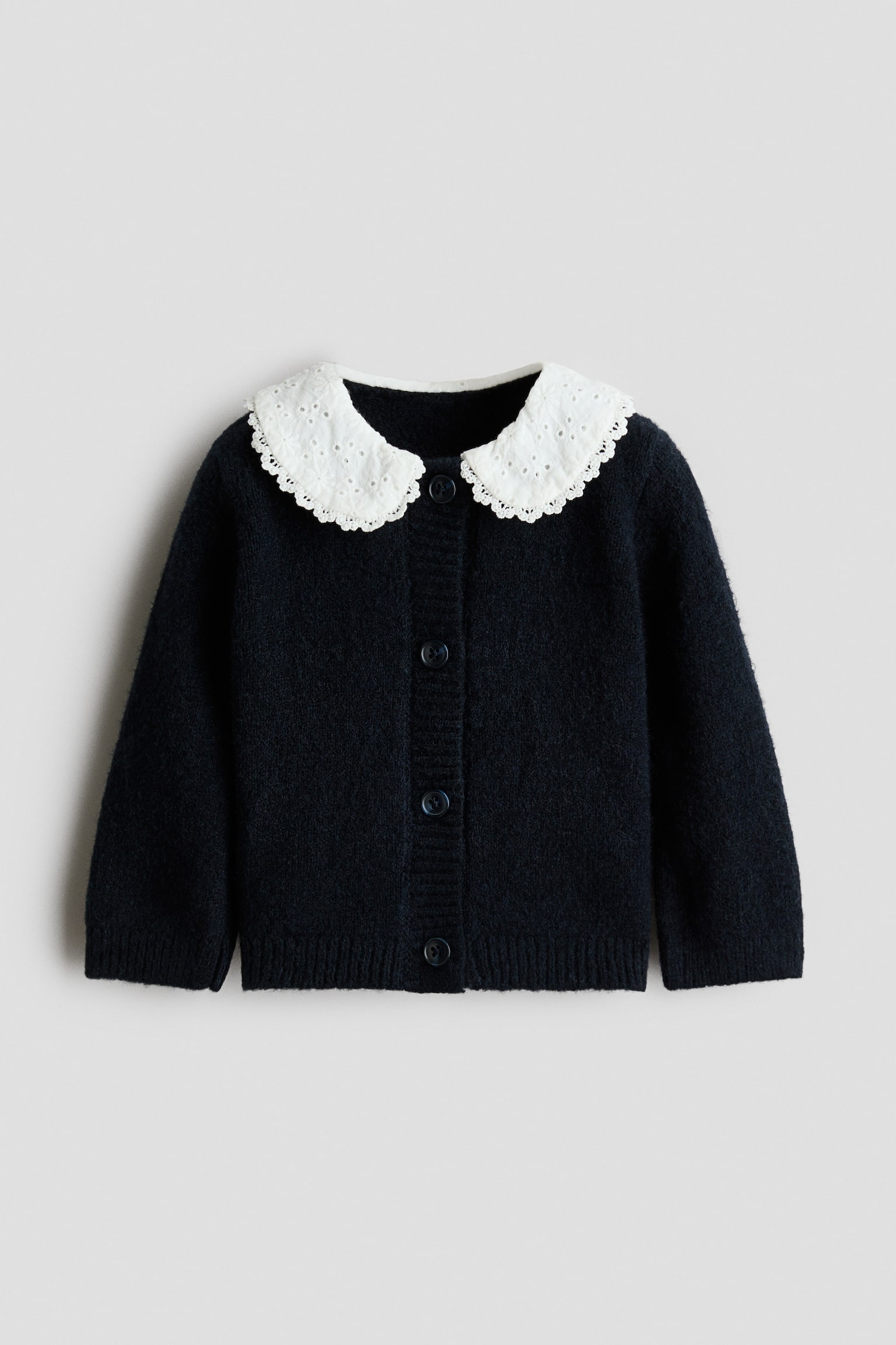 A dark navy knitted cardigan is shown from the front, featuring a contrasting white Peter Pan collar with eyelet embroidery and scalloped edges. The cardigan features a front button fastening, long sleeves with ribbed cuffs, and a ribbed hem.