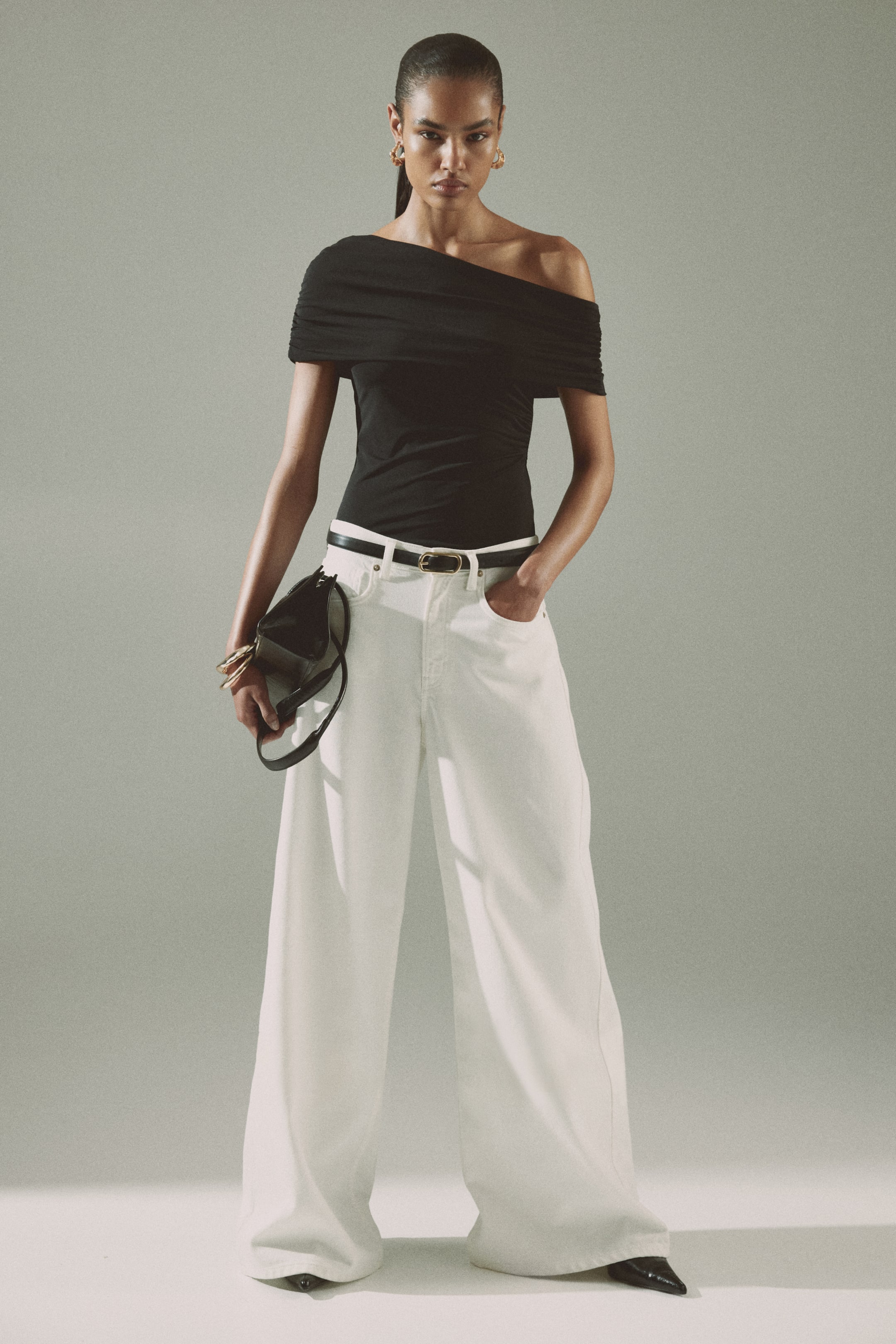 View larger image: A black top showcasing an asymmetrical, draped neckline that reveals one shoulder. The top hugs the body closely and is styled tucked into white wide-leg trousers.