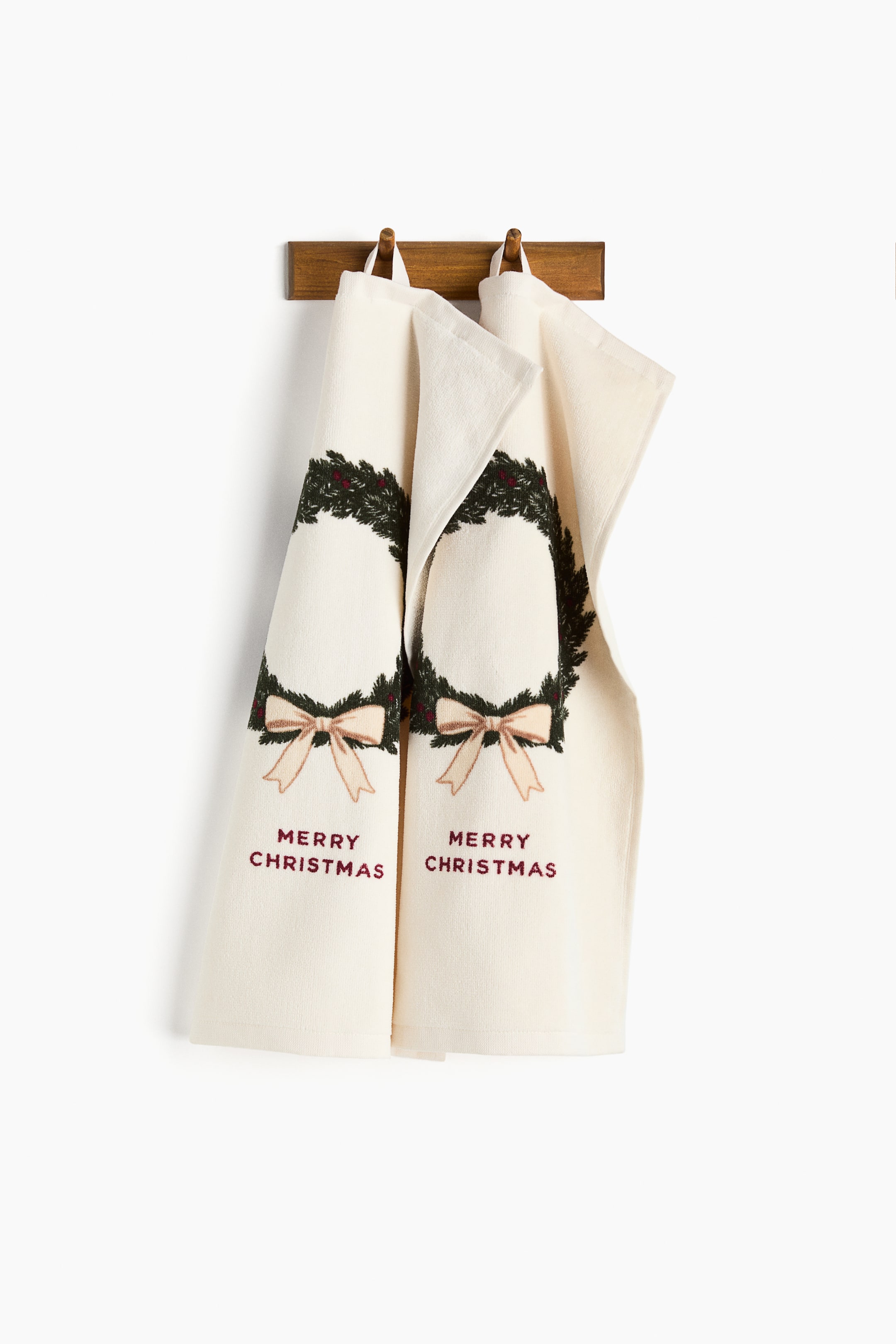 View larger image: A pair of cream soft cotton terry guest towels is hanging, with their fronts facing forward. Each towel displays a print motif of a green wreath with red berries and a light brown bow, featuring the text ‘MERRY CHRISTMAS’ below, and is finished with a fabric hanger loop on one short side.