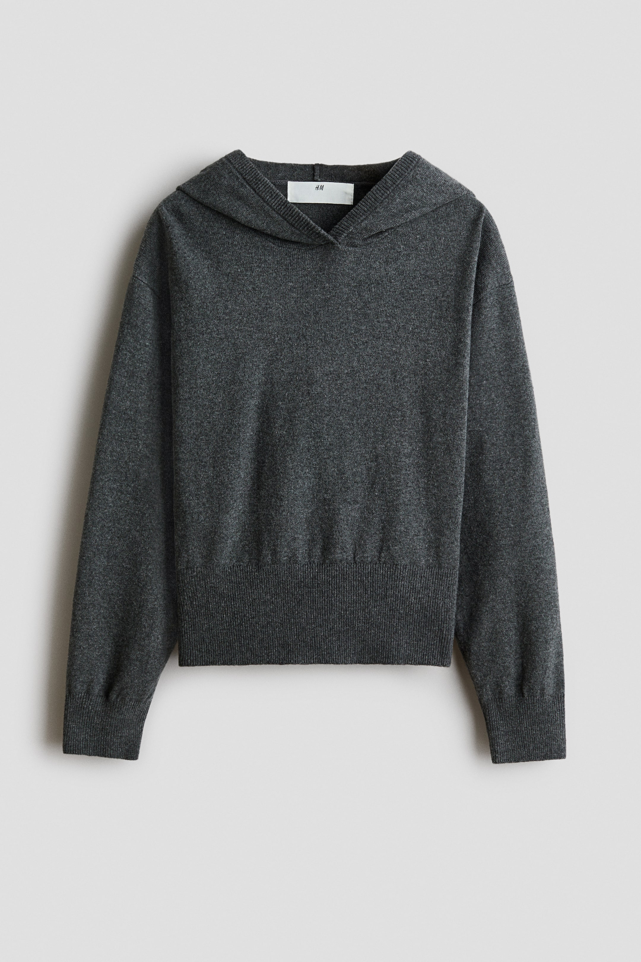 Visa större bild: En mörkgrå hoodie i finstickat material visas framifrån och har en avslappnad silhuett med korsad huva. Plagget är designat med sänkta axlar, långa ärmar med breda ribbade muddkanter och en matchande bred ribbad nederkant.