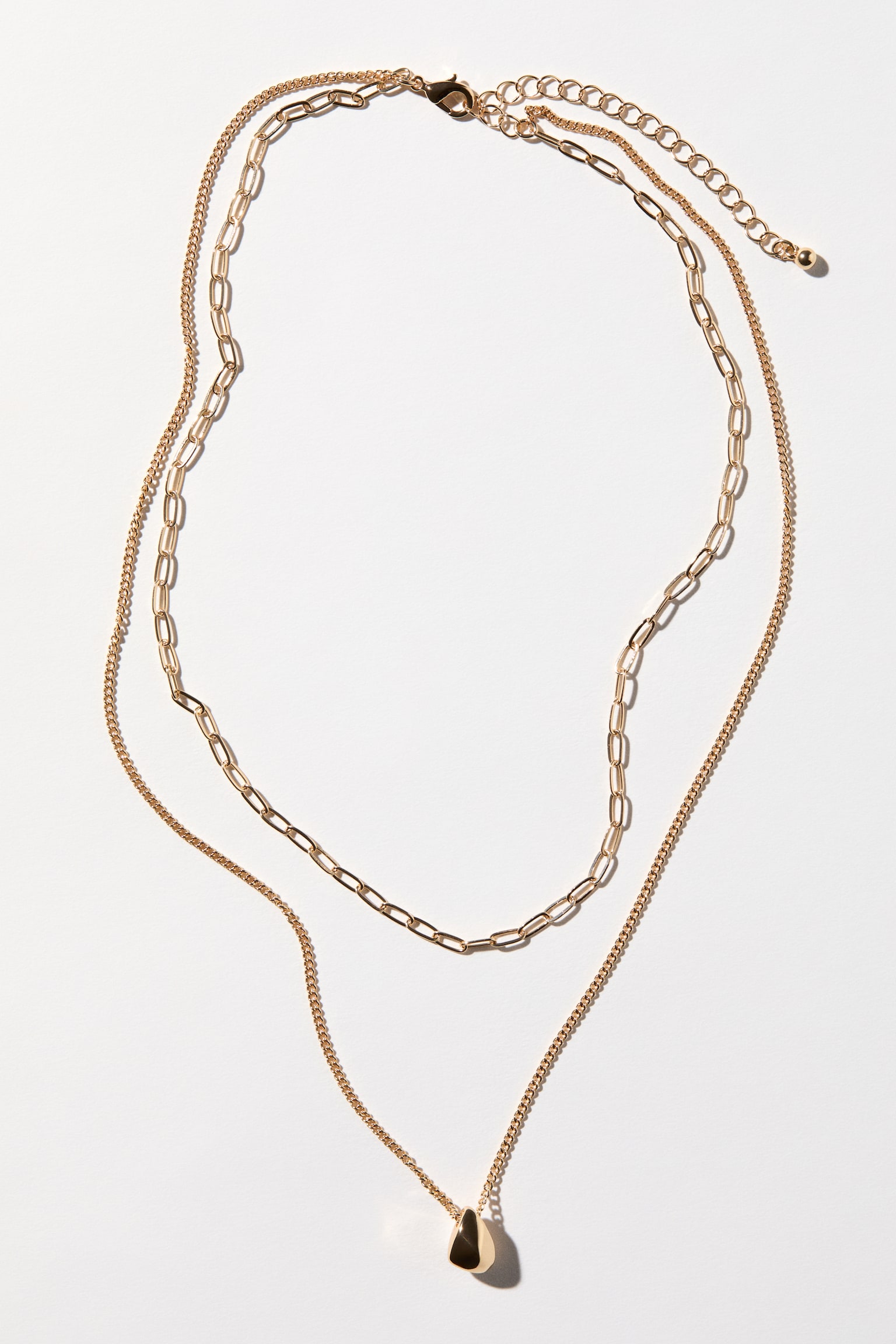 A gold-toned layered necklace lies flat, showcasing a shorter flat link chain paired with a longer, more delicate curb chain. The longer chain suspends an irregular, polished gold-toned pendant. At the back, a lobster clasp and extender chain are visible. - 1