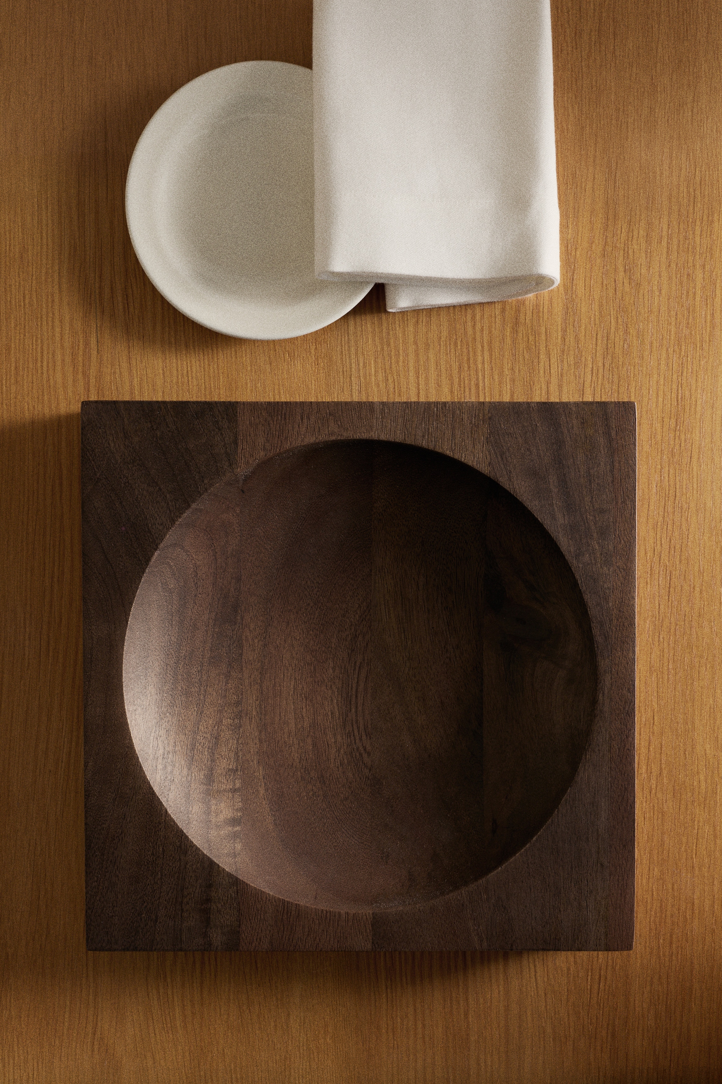 A dark brown square wooden dish displays a smooth surface with visible wood grain. A large, circular, concave well is carved into its centre.