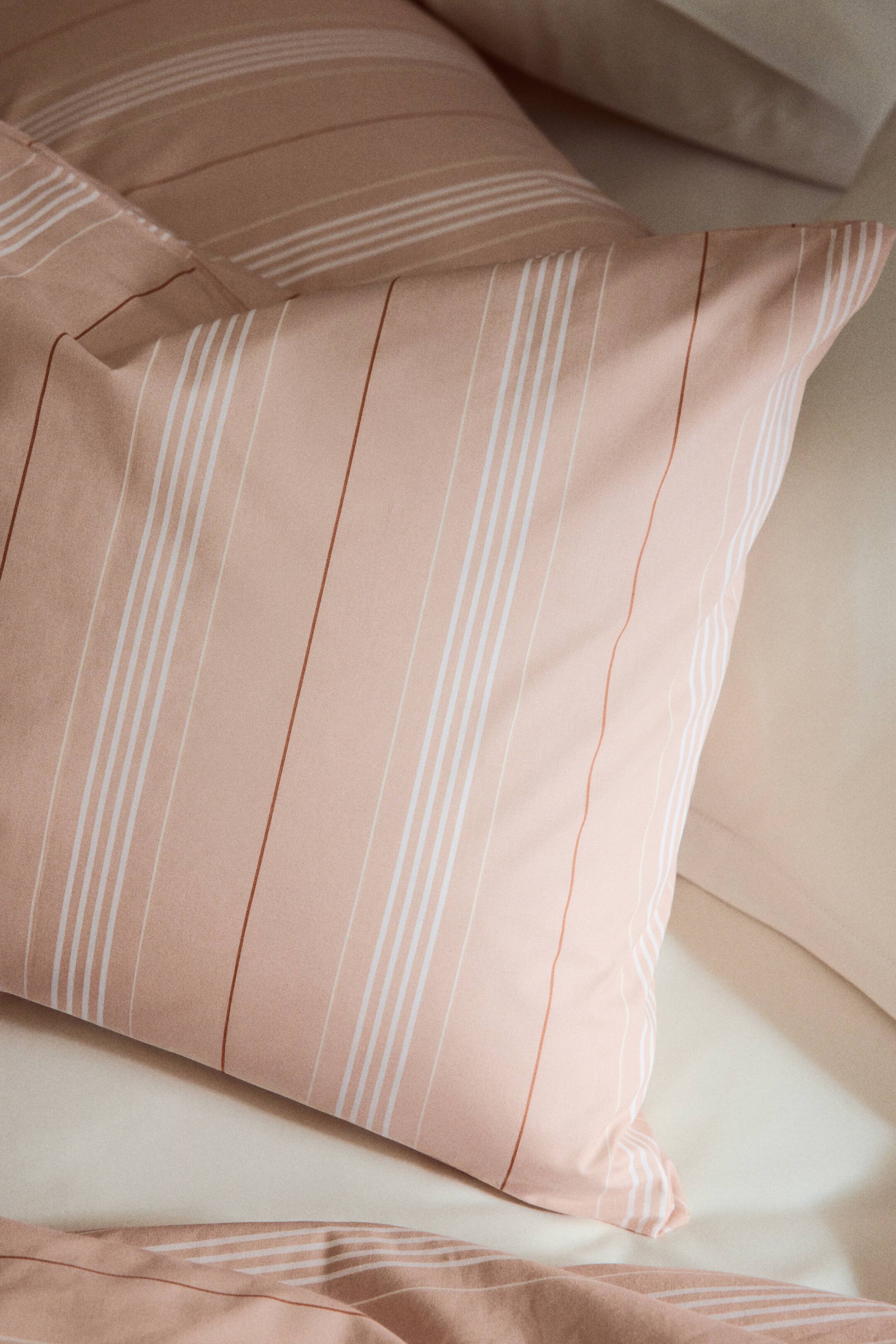 View larger image: A light pink pillowcase, showcasing vertical white, light beige, and slender brown stripes, is displayed from the front and side, angled to the right. It harmonises with a visible matching duvet cover in the foreground and a second pillowcase partially visible behind it.