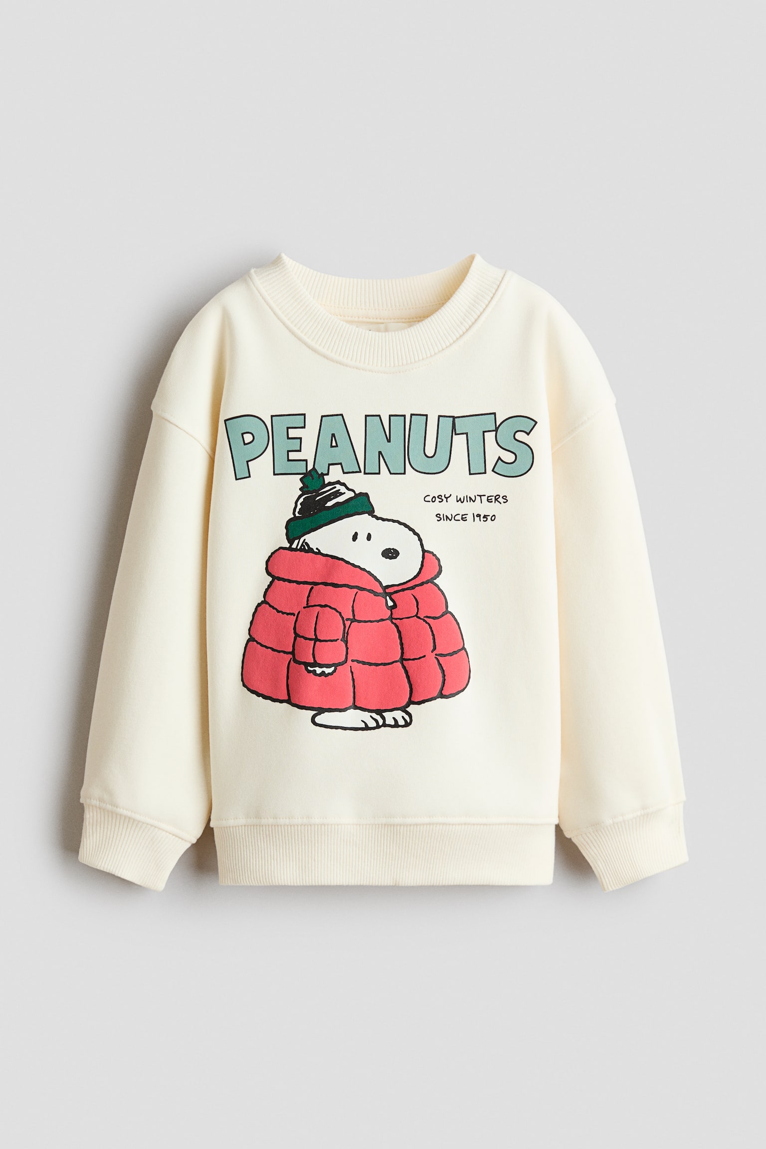 A cream-coloured sweatshirt is shown from the front, featuring a round, rib-trimmed neckline, dropped shoulders, and ribbed cuffs and hem. The front displays a printed graphic with the word "PEANUTS" in light green above an illustration of Snoopy wearing a red puffer jacket and a green beanie, alongside the text "COSY WINTERS SINCE 1950."