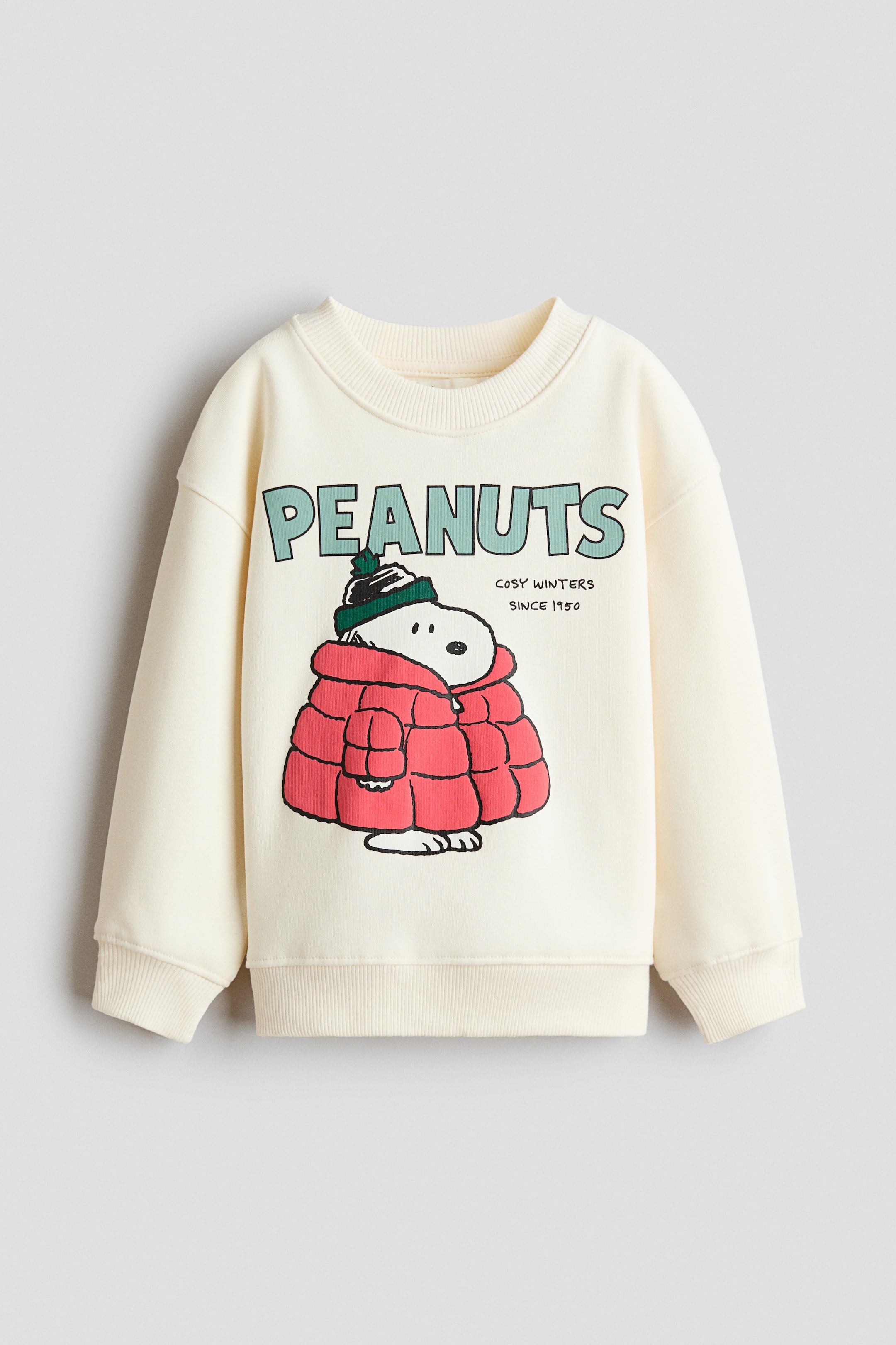 View larger image: A cream-coloured sweatshirt is shown from the front, featuring a round, rib-trimmed neckline, dropped shoulders, and ribbed cuffs and hem. The front displays a printed graphic with the word "PEANUTS" in light green above an illustration of Snoopy wearing a red puffer jacket and a green beanie, alongside the text "COSY WINTERS SINCE 1950."