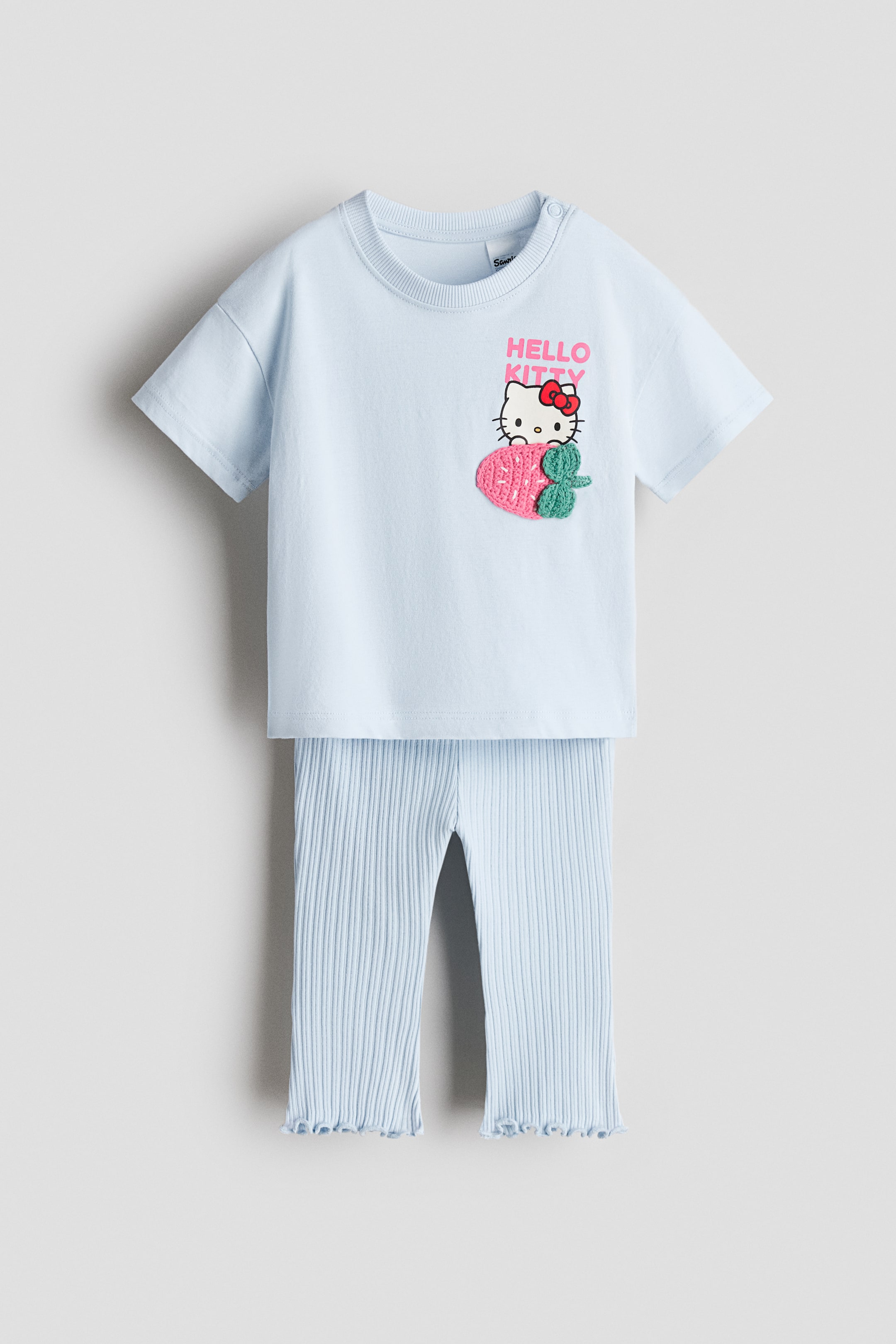 Ver imagem maior: Um conjunto de duas peças azul-claro apresentado de frente, composto por uma t-shirt de mangas curtas e calças caneladas a condizer. A t-shirt apresenta uma gola redonda canelada, fecho de mola no ombro e um motivo "Hello Kitty" com um apliqué de morango em croché, enquanto as calças apresentam bainhas com folhos.