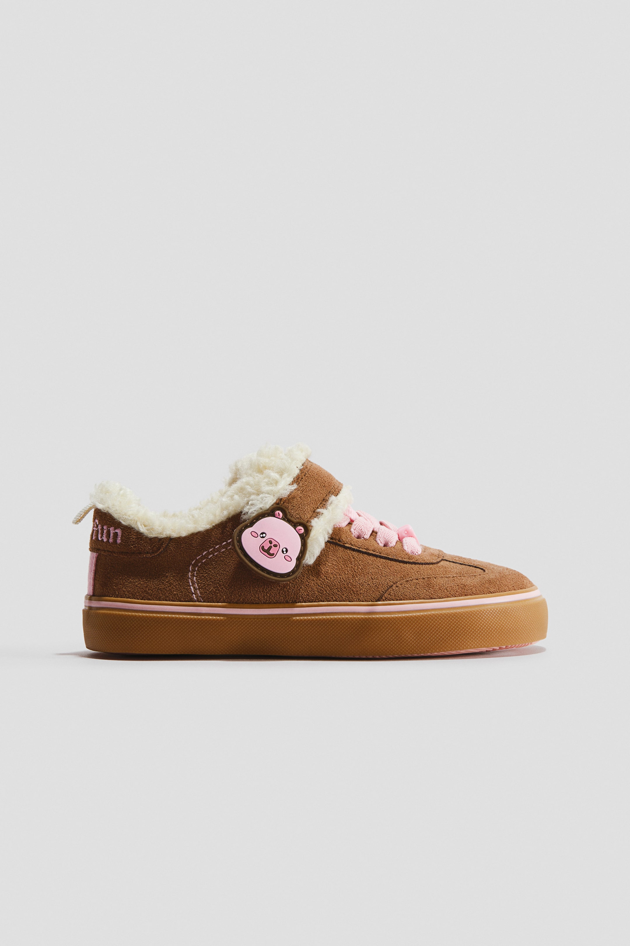Agrandir l'image: Cette sneaker en daim marron est présentée de profil, avec une doublure en fausse peau lainée crème le long du col et de la languette. Elle est dotée de lacets roses, d’une bride auto-agrippante ornée d’une breloque rose en forme d’animal de dessin animé, ainsi que d’une semelle extérieure en caoutchouc couleur gomme avec une fine bande rose.