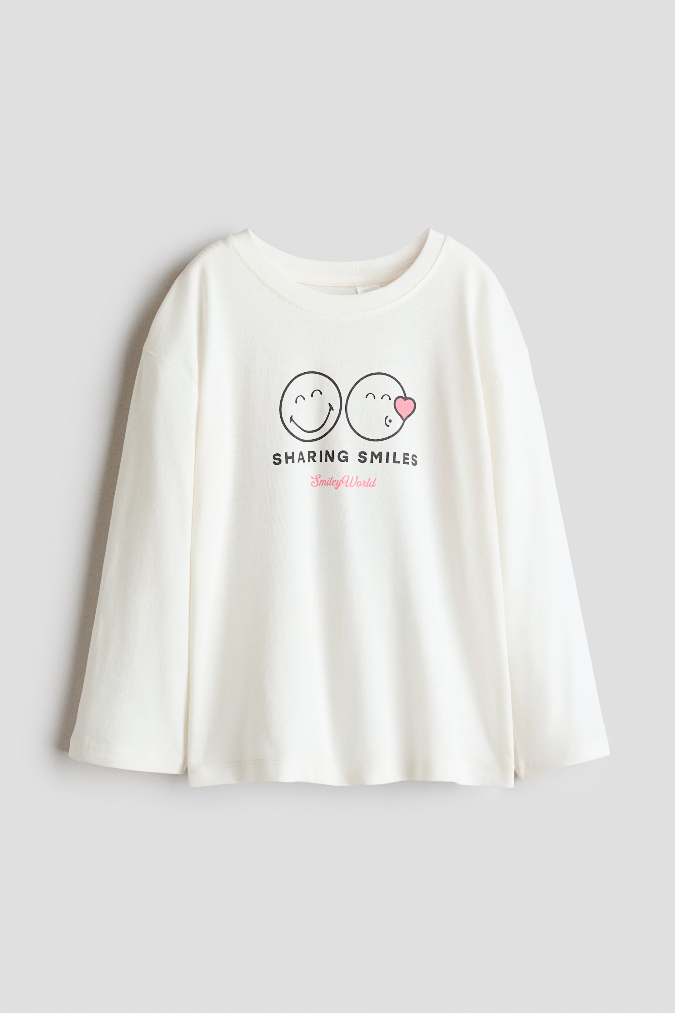 View larger image: An off-white long-sleeved top is shown facing forward, featuring a crew neck, relaxed fit, and a straight hem with small side splits. The front displays a graphic of two circular emoji faces, one smiling and one blowing a kiss with a pink heart, with the text "SHARING SMILES" appearing below them, and "Smiley World" in pink script below that.