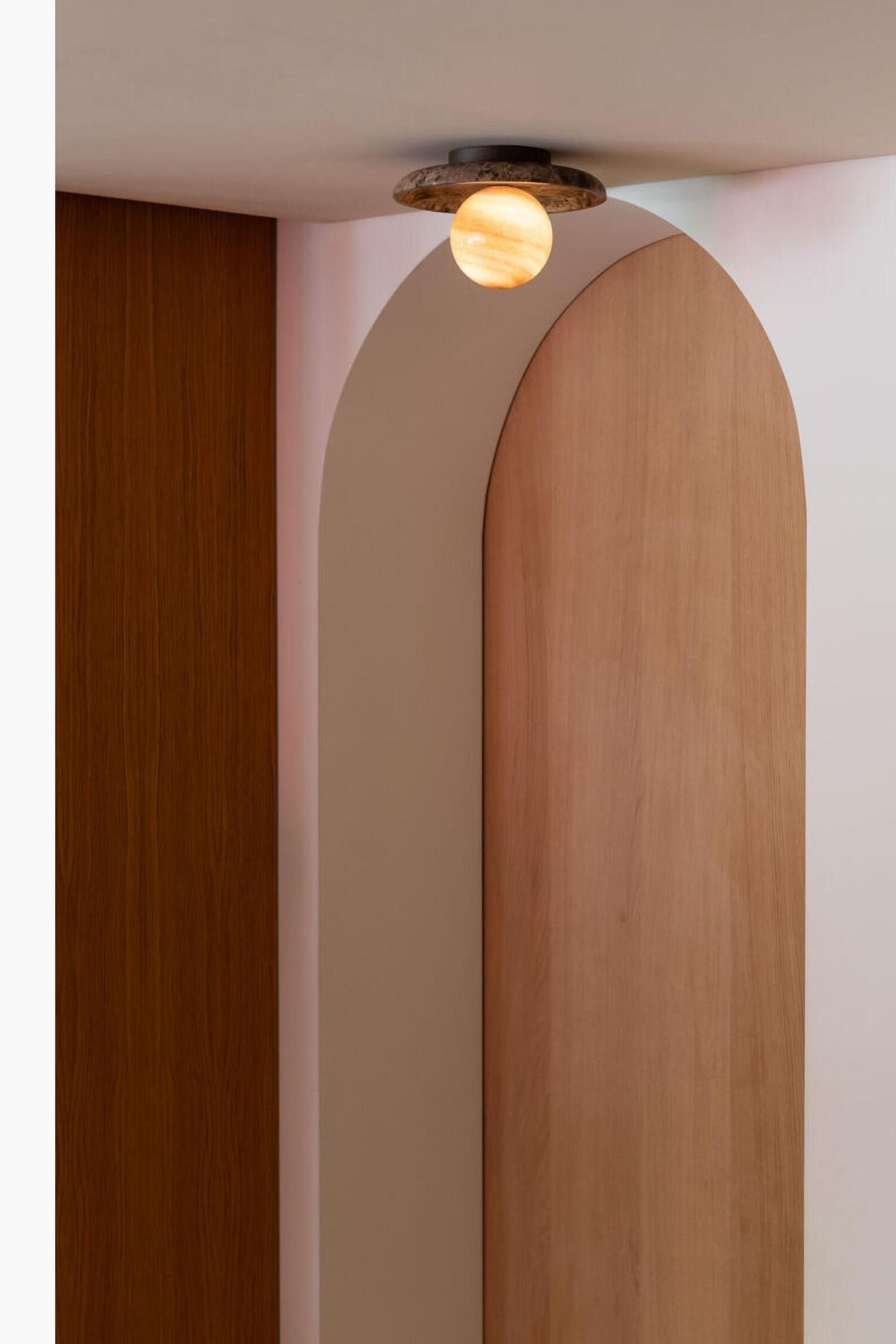 Een opbouwarmatuur toont een gloeiende, bolvormige kap met warme gele en oranje aders. De verlichte bol steekt uit vanuit het midden van een brede, holle, cirkelvormige plafondkap, vervaardigd uit bruin gemarmerde steen.