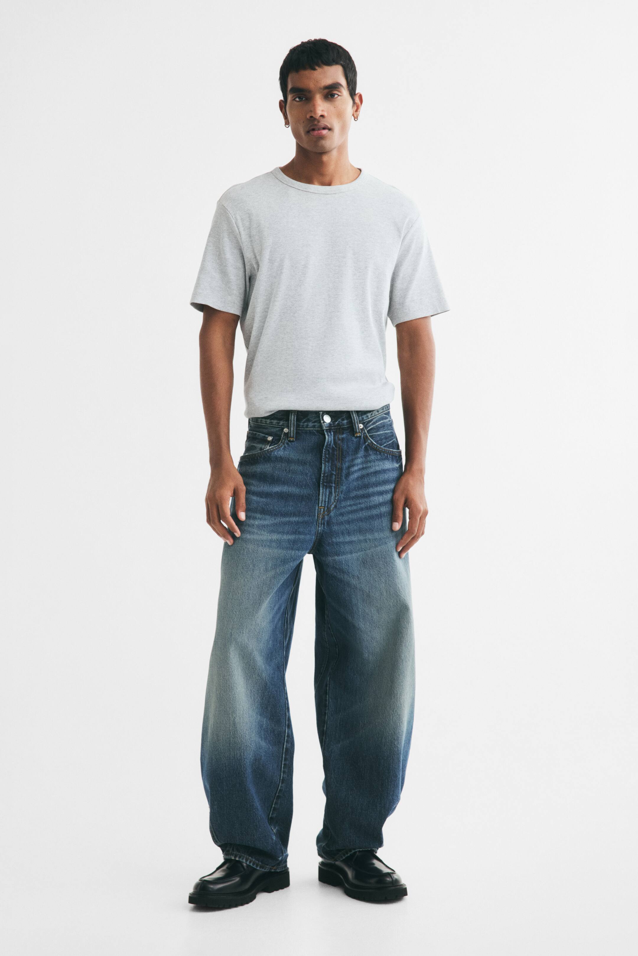 Vis større bilde: Blå vide jeans i denim med falmet vask bæres av modellen. En grå kortermet T-skjorte med rund hals er stukket ned i jeansen, og det brukes svarte sko med kraftig såle.