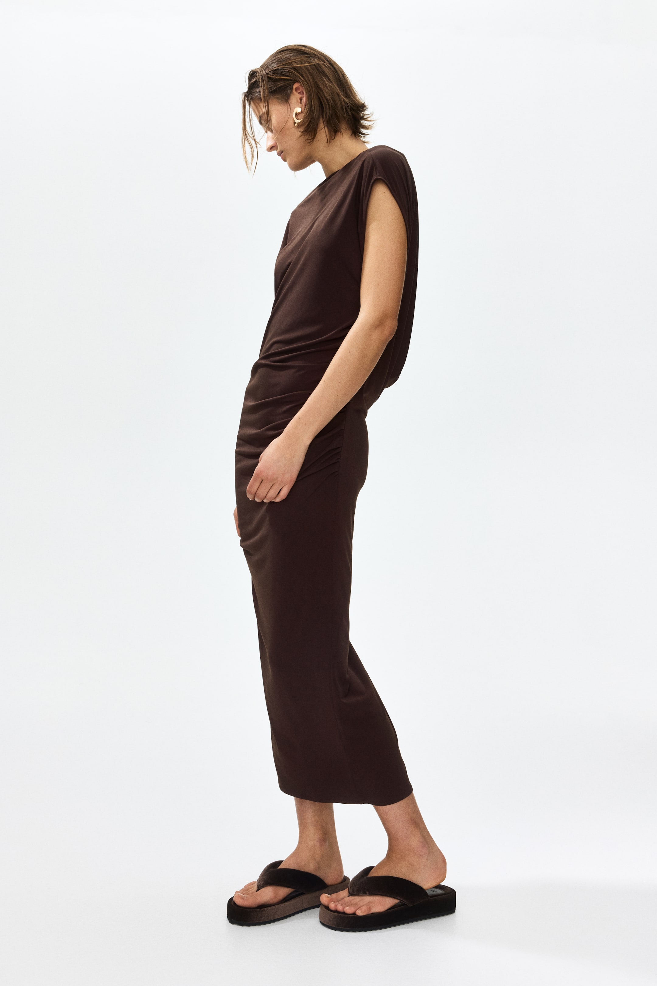 Grösseres Bild ansehen: Ein dunkelbraunes Midikleid wird von der Seite präsentiert, mit einem runden Ausschnitt, kurzen Ärmeln und gerafften Details entlang der Seitennähte, die eine drapierte Silhouette schaffen.
