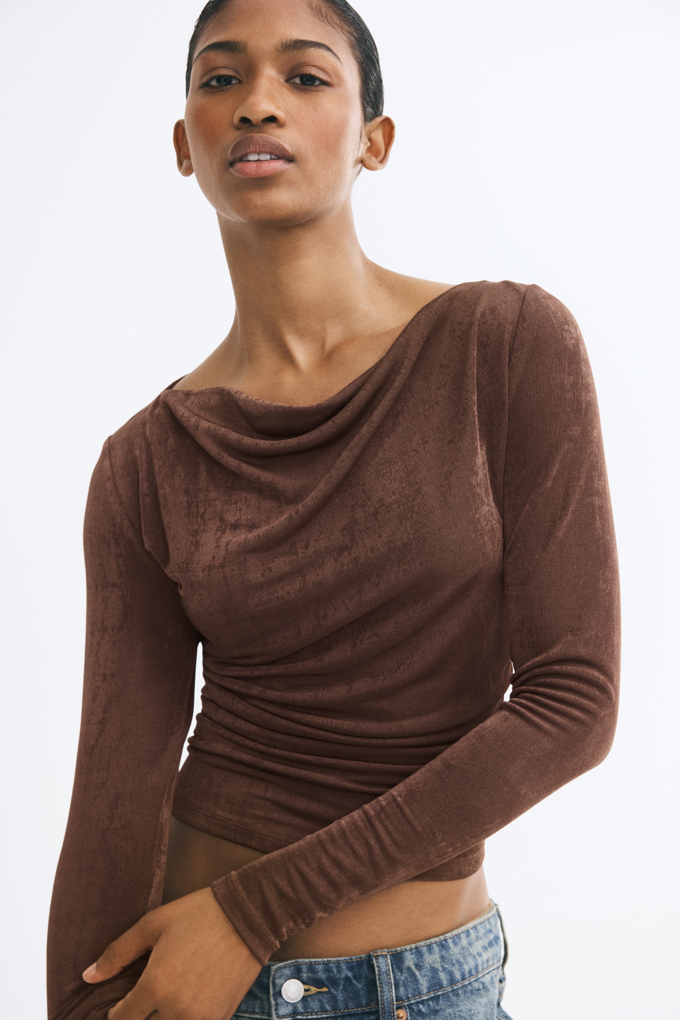 A brown, textured-fabric long-sleeved top is shown from the front, featuring a draped cowl neckline and ruched side seams that create horizontal gathers across the lower torso.