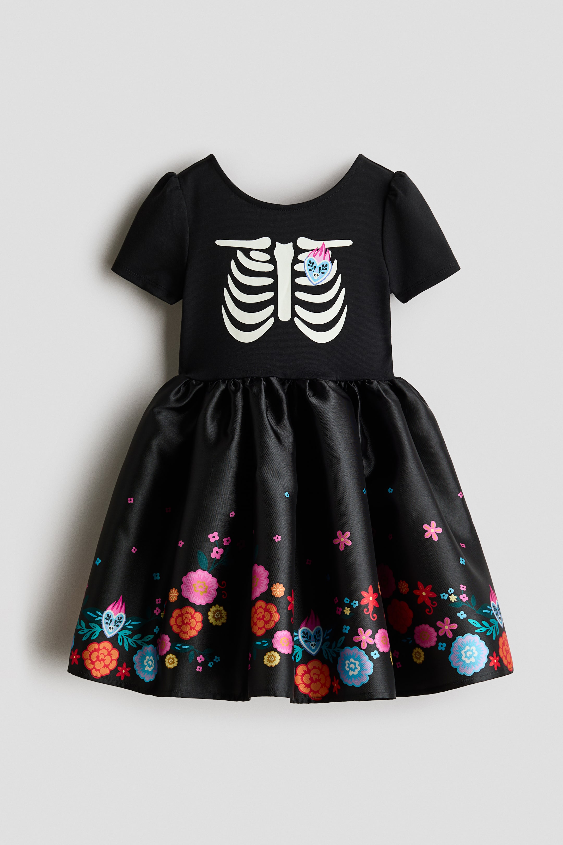 Ampliar la imagen: Un vestido negro de frente presenta mangas cortas ligeramente abullonadas y un escote redondo. Su corpiño muestra un gráfico de costillas de esqueleto blanco con un corazón bordado colorido en el lado izquierdo, mientras que la falda recogida exhibe un borde de patrones florales y de corazones vibrantes a lo largo del dobladillo.