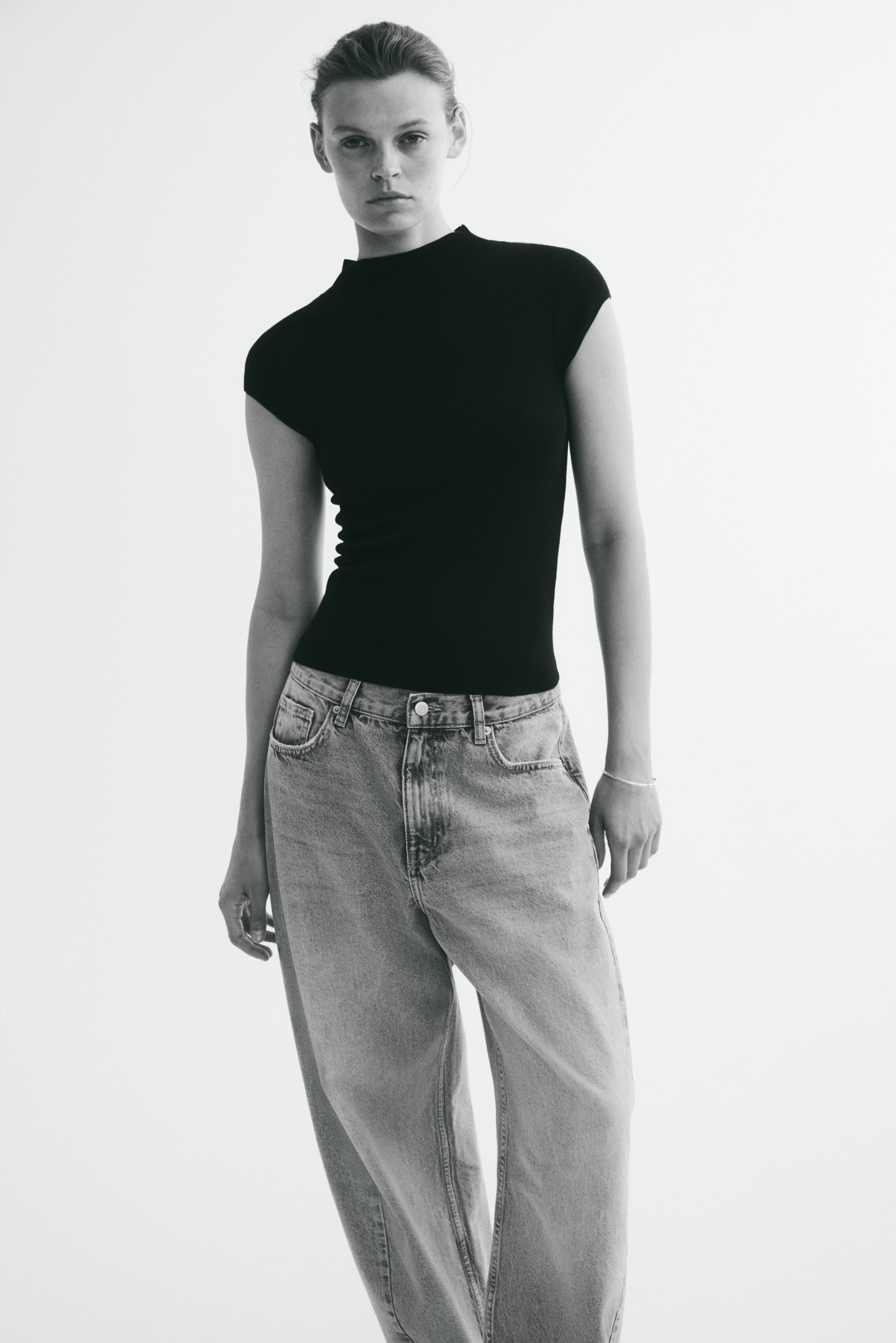 View larger image: A fitted black top in a fine knit is being worn by the model, featuring a high neck, cap sleeves, and a straight hem. The top is styled with loose-fitting, light-wash denim jeans.