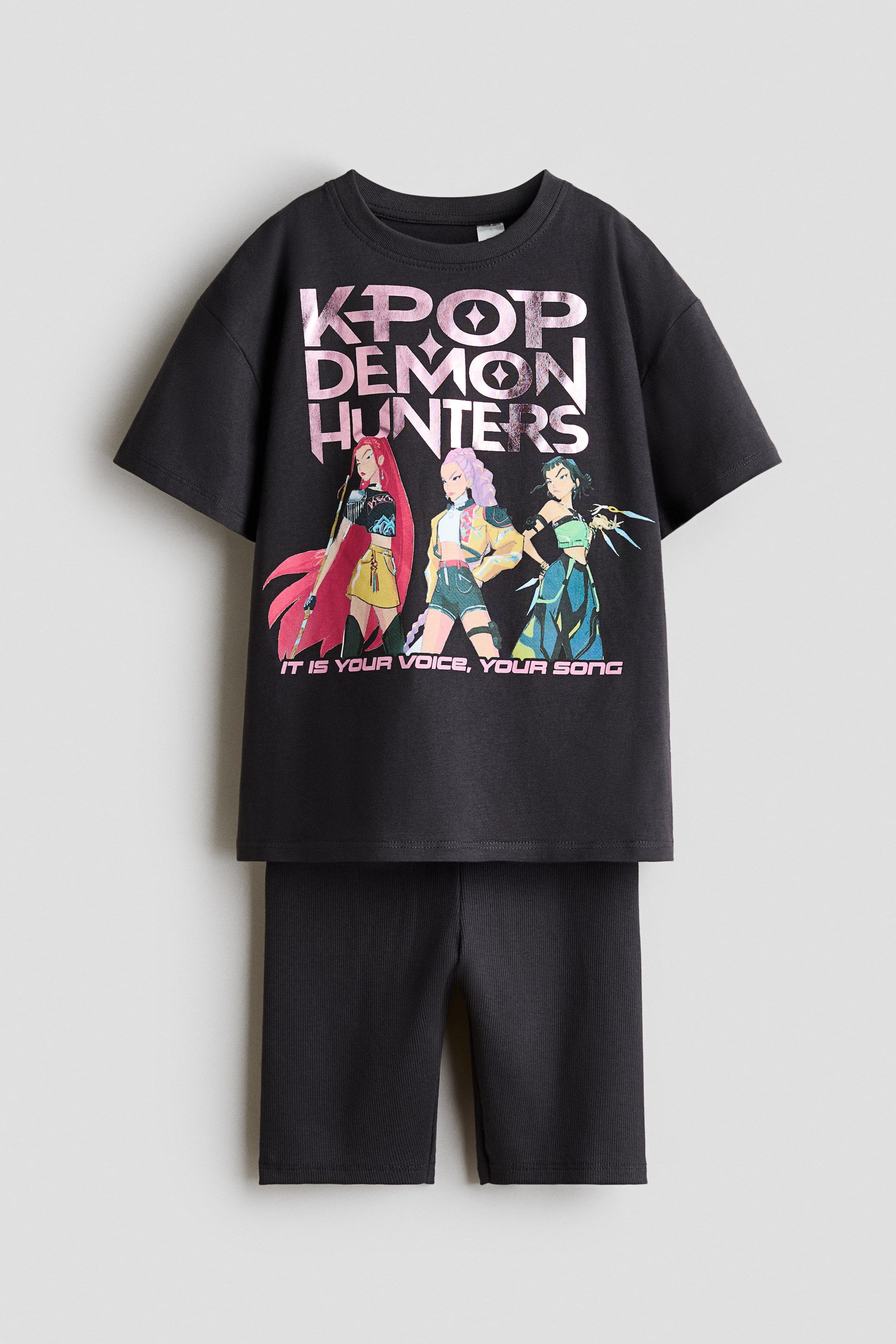 Visa större bild: Ett mörkgrått tryckt set bestående av en T-shirt och matchande ribbade cykelshorts, presenterat framifrån. T-shirten visar "KPOP DEMON HUNTERS" i metalliskt rosa text ovanför tre illustrerade kvinnliga karaktärer och "IT IS YOUR VOICE, YOUR SONG" nedanför, med en ribbad halsringning och nedhasade axlar. Cykelshortsen är designade med en elastisk midja.