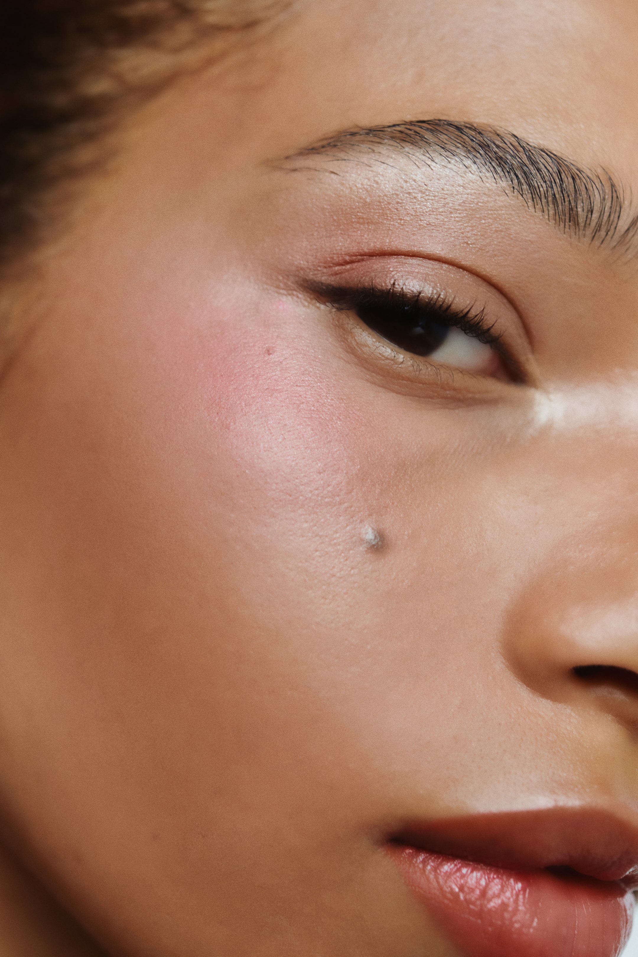 Größeres Bild anzeigen: Auf der rechten Gesichtshälfte wird Make-up aufgetragen, das ein dezentes rosa Lidschatten auf dem Augenlid zeigt, zusammen mit einem dünnen schwarzen Winged Eyeliner und Mascara auf den oberen Wimpern. Ein sanfter rosiger Rouge hebt den Wangenknochen hervor, und ein glänzendes rötlich-braunes Produkt bedeckt die Lippen.