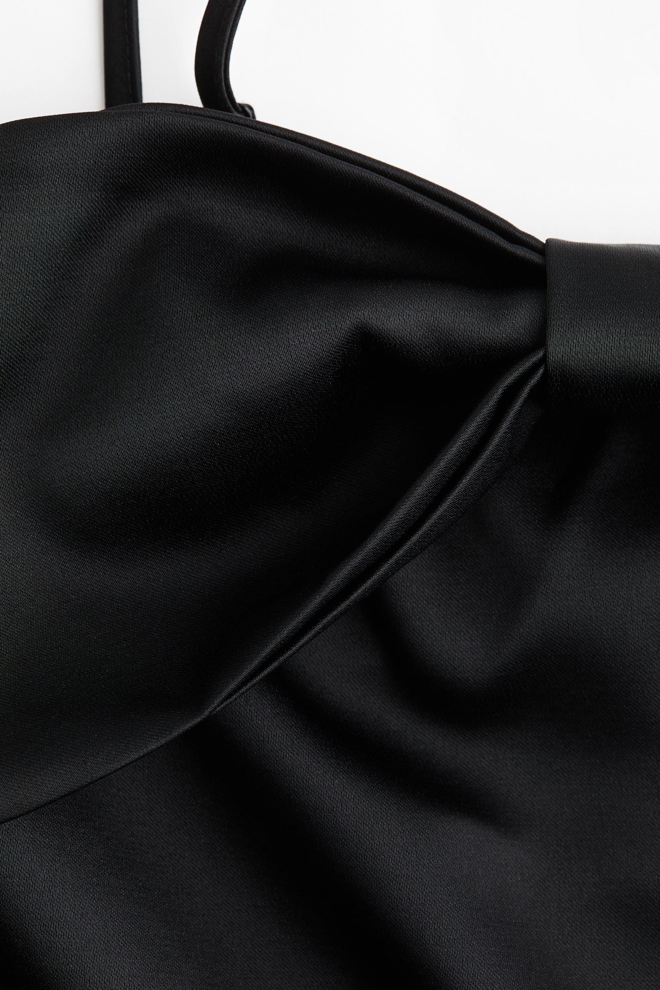 Agrandir l'image: Un vêtement noir présente un grand nœud structuré sur le devant, positionné vers la droite. Le tissu lustré tombe avec fluidité et forme de douces plis autour du nœud central, et une fine bretelle est visible en haut à gauche.