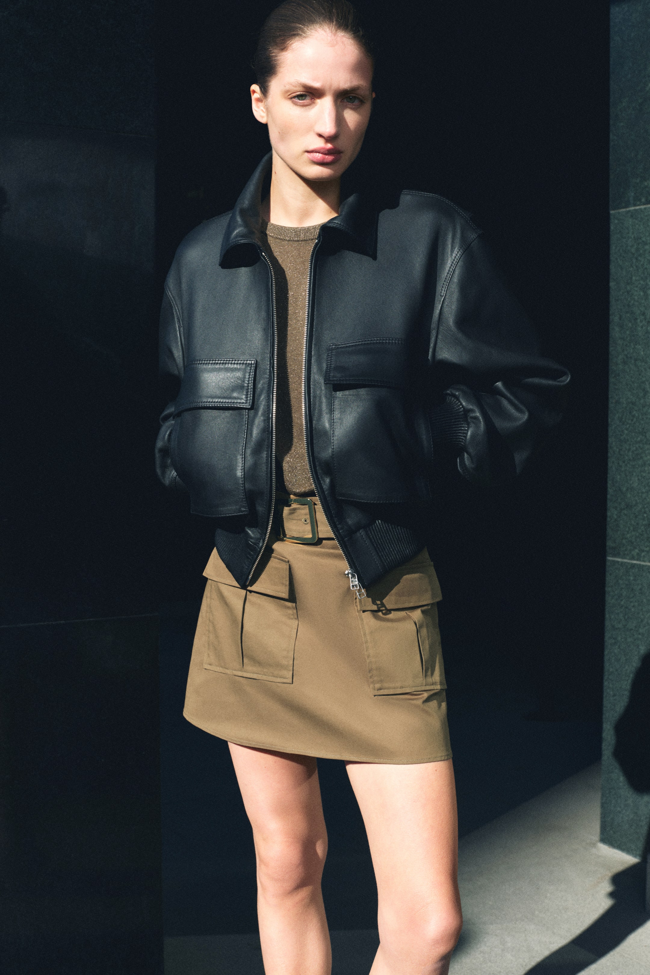 הצגת תמונה גדולה יותר: מעיל בומבר מעור שחור מוצג, עם צווארון רחב, סגירת רוכסן מלאה ושני כיסי דש במותן, יחד עם חפתים ומכפלת מצולעים. המעיל משולב מעל חולצת סריג זוהרת בצבע זית, שמשולבת עם חצאית מיני בצבע חאקי המציגה חגורה עם אבזם מרובע ושני כיסי טלאי קדמיים.