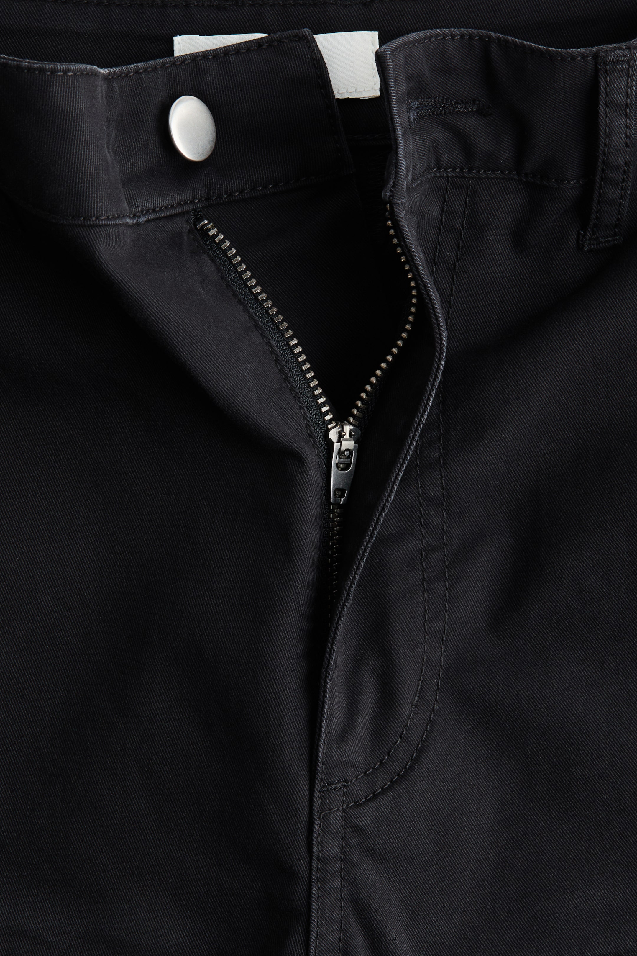 Xem ảnh lớn hơn: Quần đen có một cái nhìn phía trước về phần đóng, với một nút bấm tông màu bạc gắn chặt cạp quần. Một khóa kéo tông màu bạc được kéo một phần, lộ ra các răng khóa và đầu kéo, và vải có một họa tiết dệt chéo tinh tế với chi tiết đường chỉ may cùng màu.