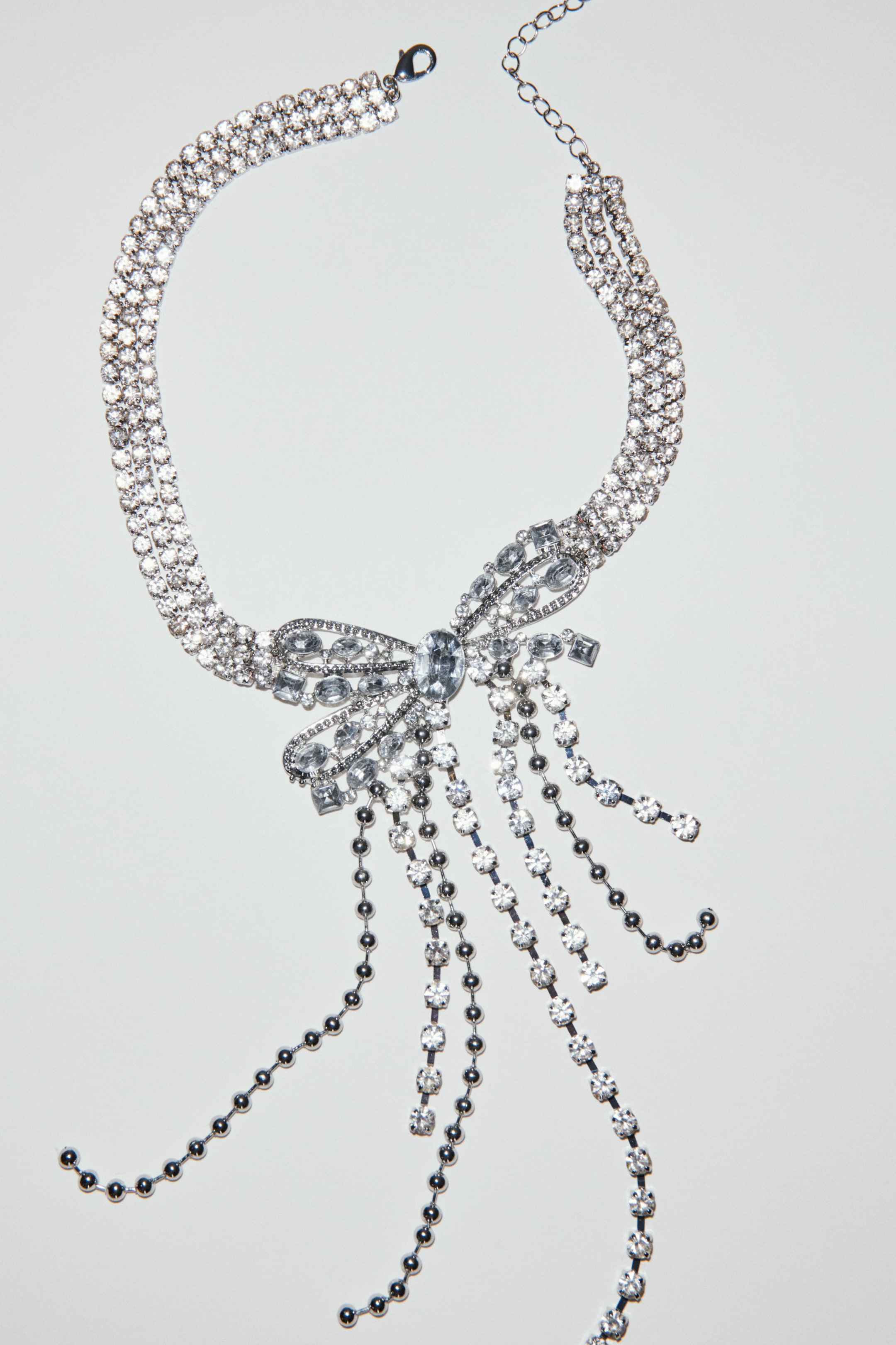 View larger image: A silver-toned necklace is presenting a triple-strand choker band, which is encrusted with clear rhinestones. A large bow-shaped pendant, adorned with various cut clear rhinestones, is positioned at its center front, from which multiple strands of dangling silver-toned chains are extending, some featuring clear rhinestones and others small silver beads. A lobster clasp and extender chain are visible at the back of the choker.