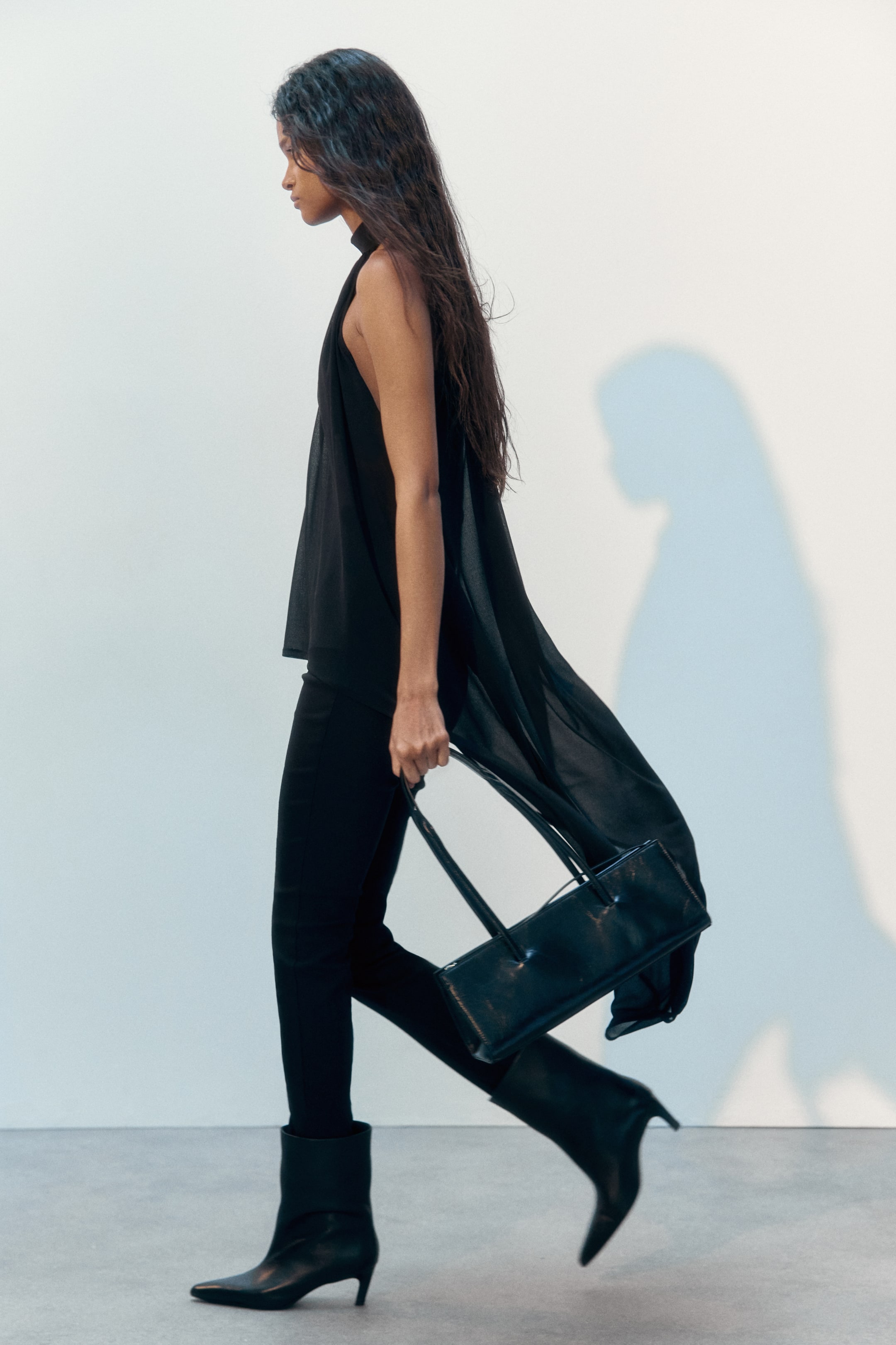 Grotere afbeelding bekijken: Een losvallende haltertop van doorschijnende jersey wordt gedragen door het model. Het heeft een opstaande kraag en plooien die een subtiel gedrapeerd effect creëren, met een asymmetrische zoom die meebeweegt terwijl het model loopt.