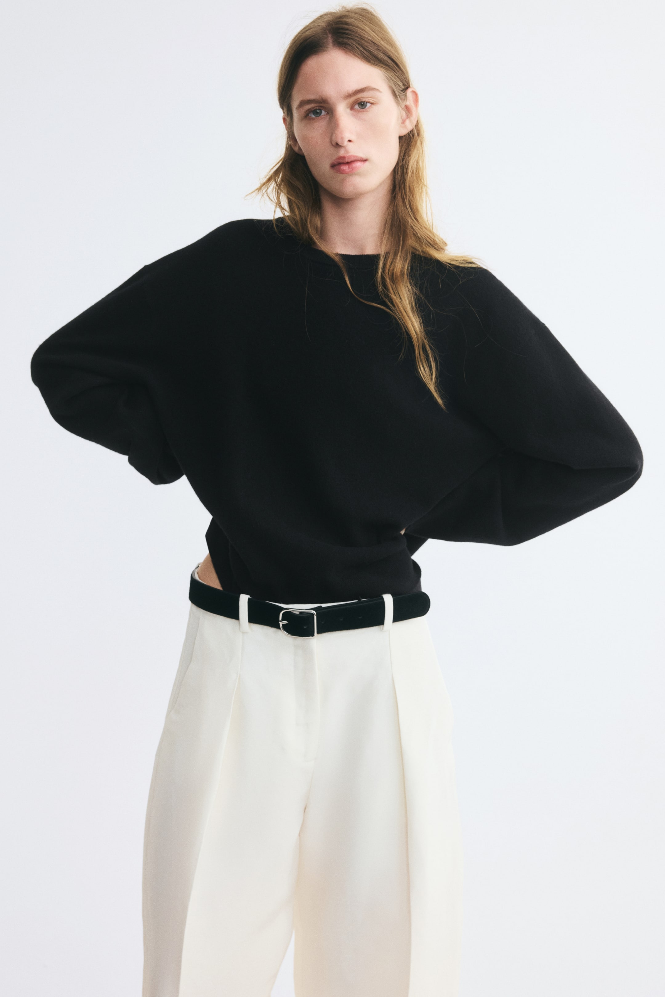 Grotere afbeelding bekijken: Een oversized zwart gebreid wollen sweater wordt gedragen door het model, met een ronde hals, verlaagde schouders en lange mouwen. De sweater is ingestopt in een witte broek, vastgezet met een zwarte riem, terwijl het model poseert met de handen in de heupen.