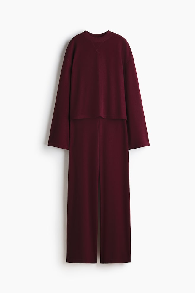 A maroon two-piece set is presented from the FRONT, featuring a long-sleeved, crew neck top with a V-stitch detail below the neckline and a relaxed fit. The top's straight hem is overlapping matching full-length, wide-leg pants that are showcasing a central seam running down the front of each leg. - 1